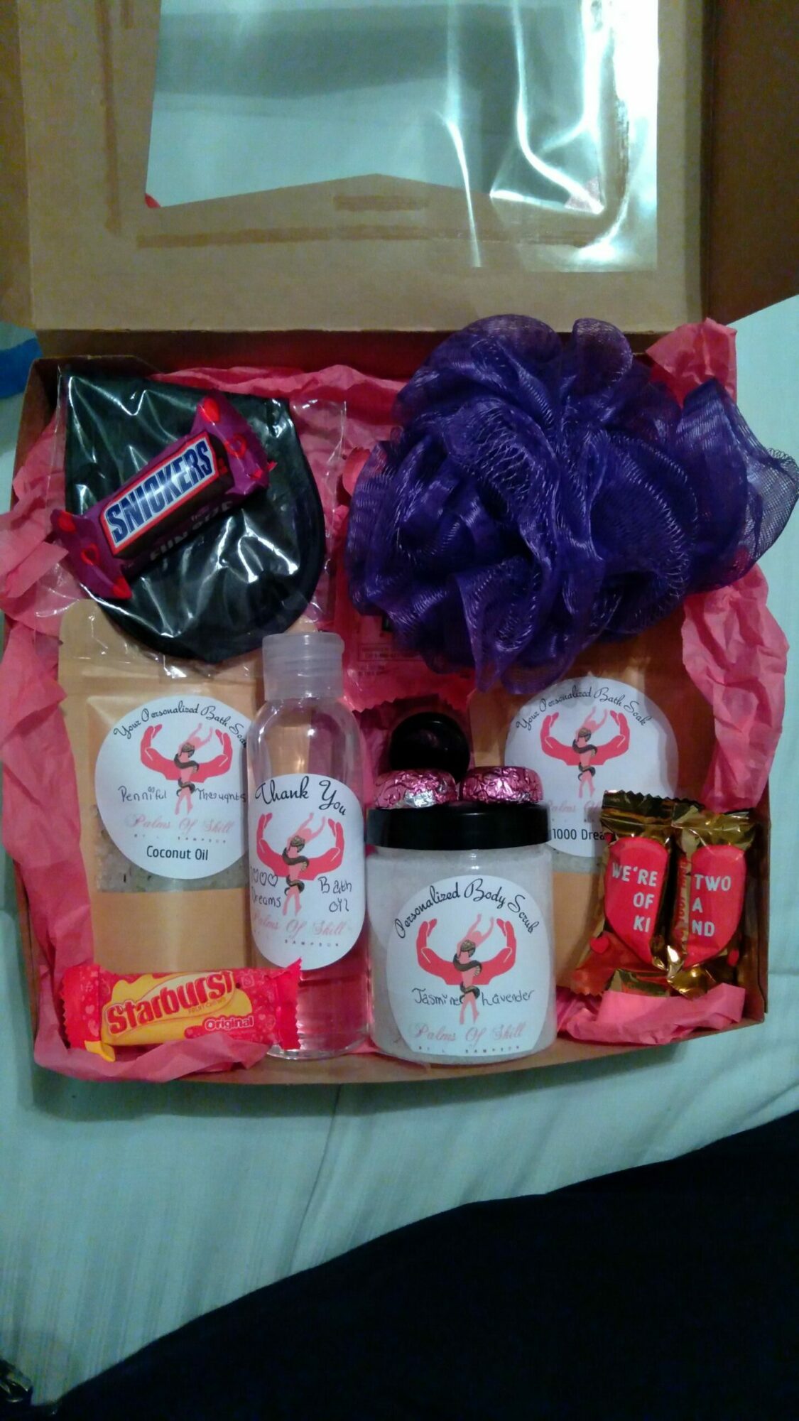 Gift box with bath products, a purple bath sponge, and small packaged items inside a pink wrapping paper, on a bed.