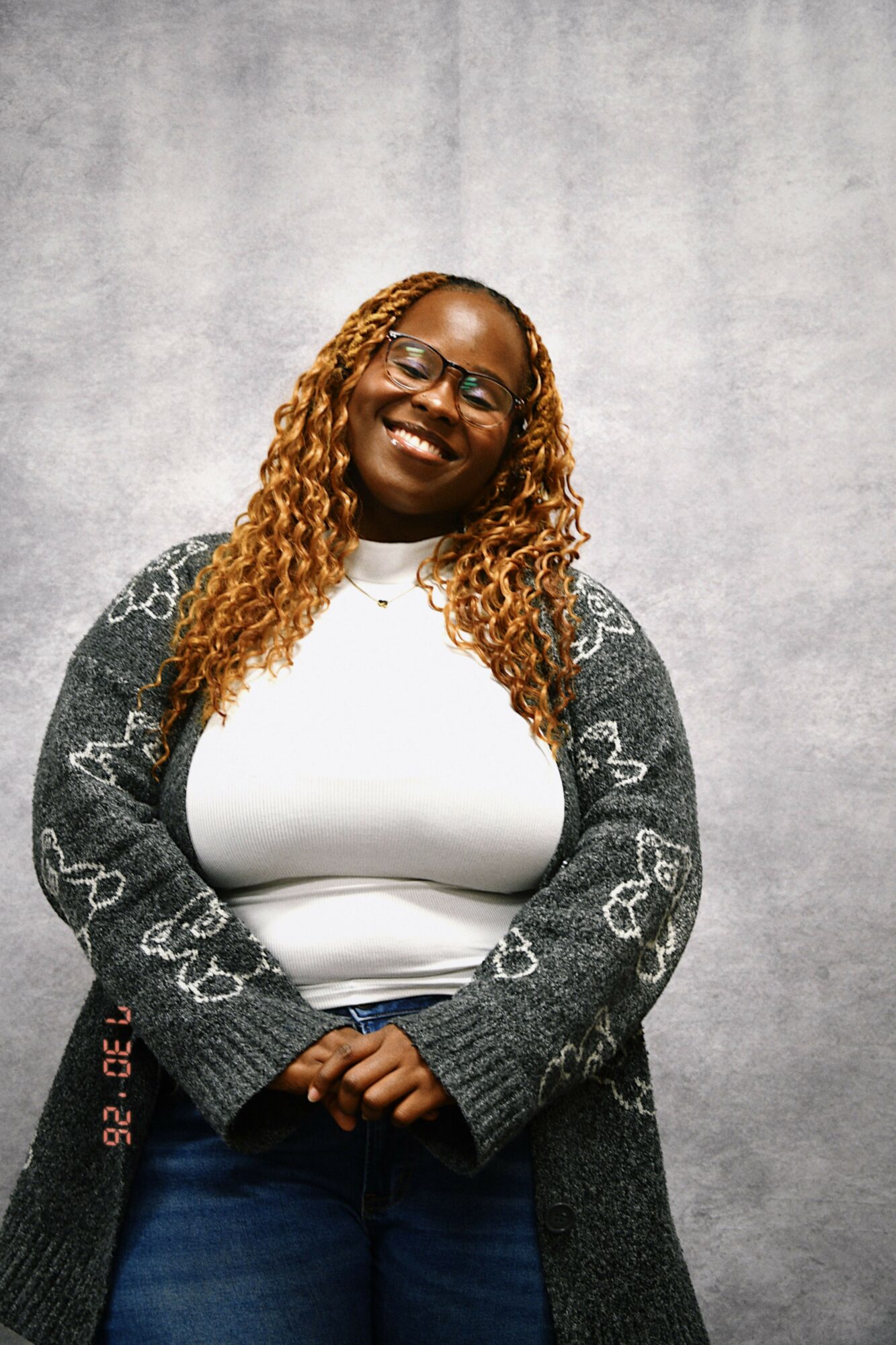 Woman with curly hair and glasses smiling, wearing a white top and gray cardigan, standing against a gray background.
