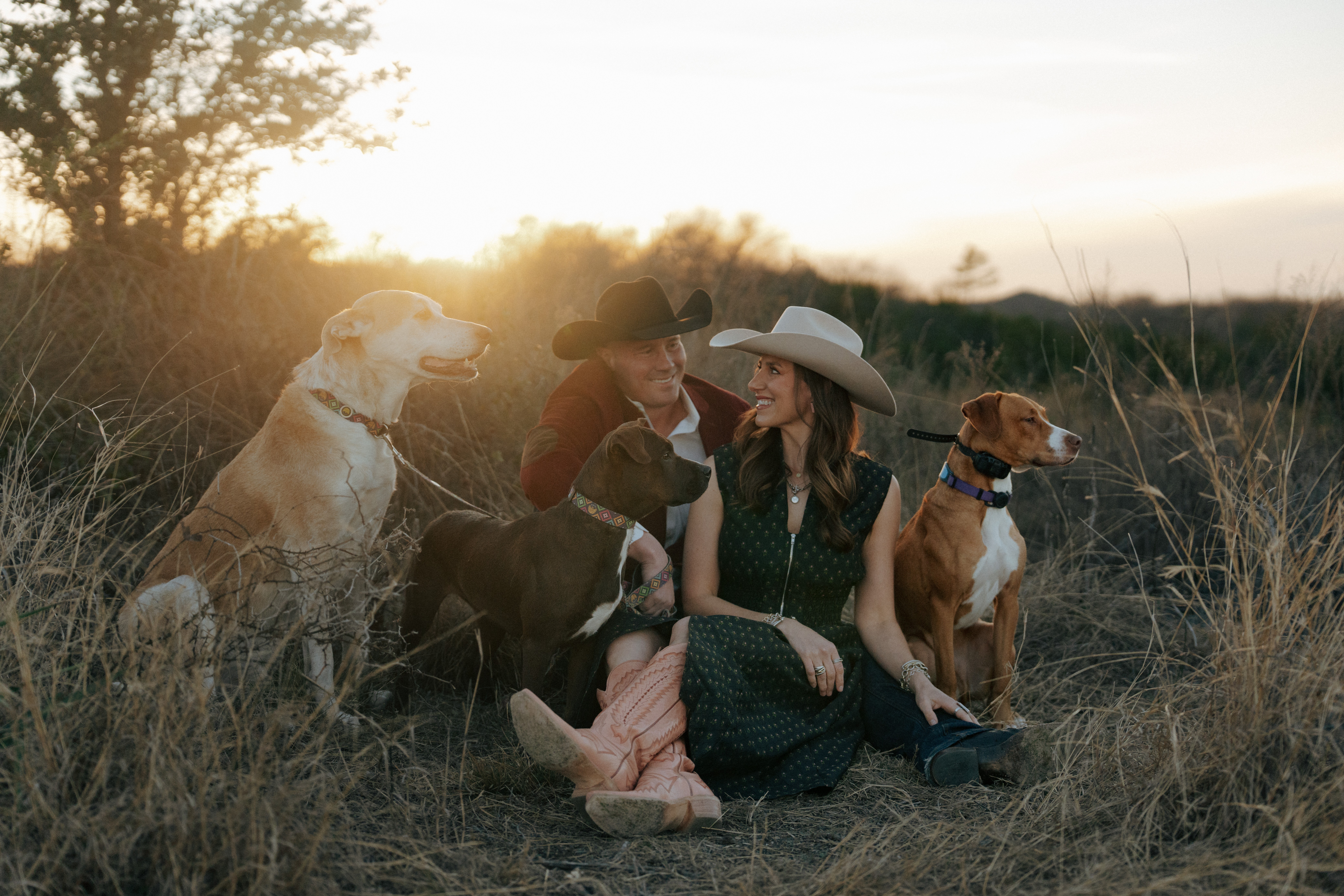 Two people sitting outdoors with four dogs during sunset, wearing hats and casual clothing, in a grassy field.