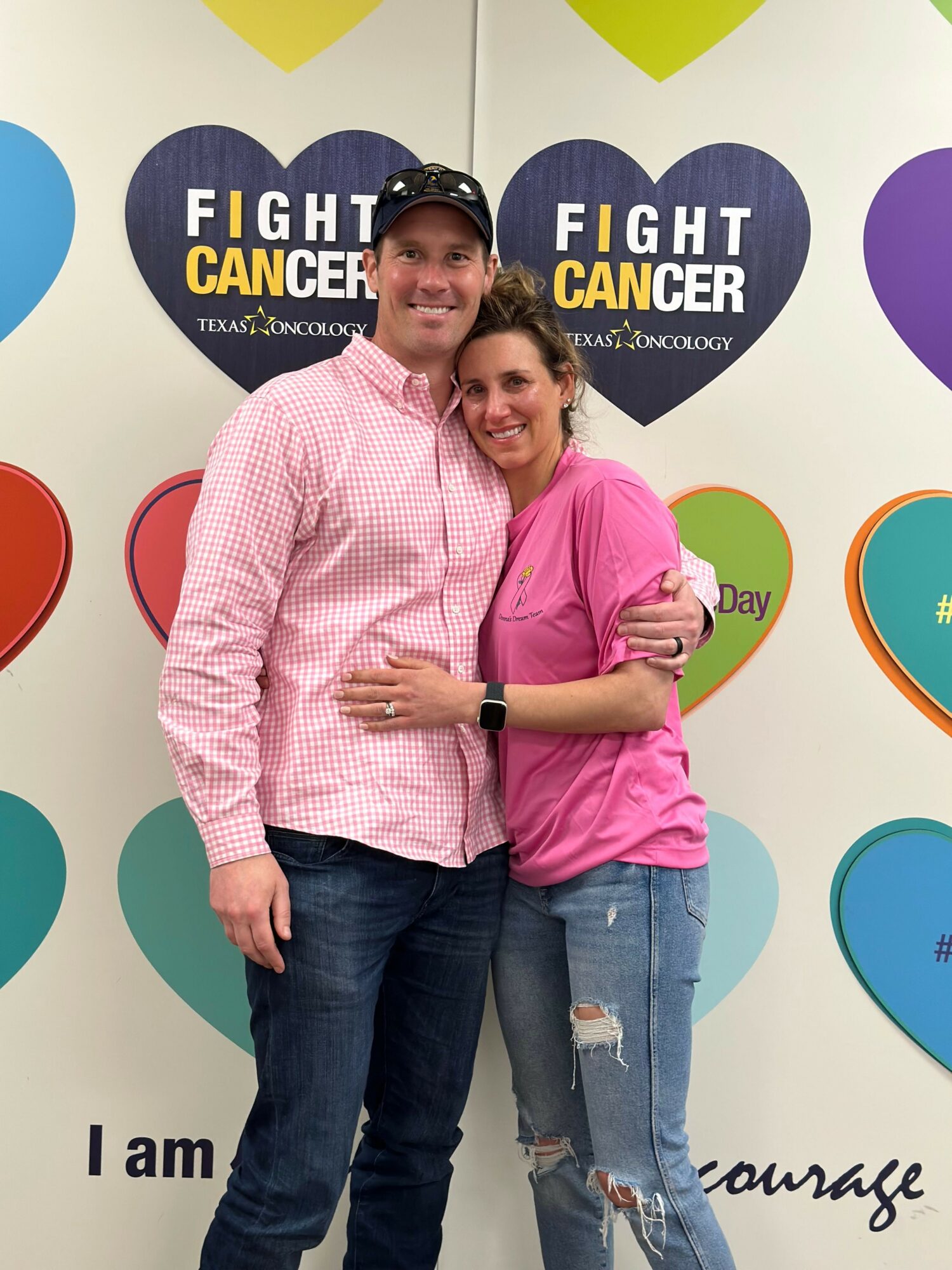 Two people standing in front of a colorful wall with cancer awareness messages, one wearing a pink shirt and the other in a pink checkered shirt.