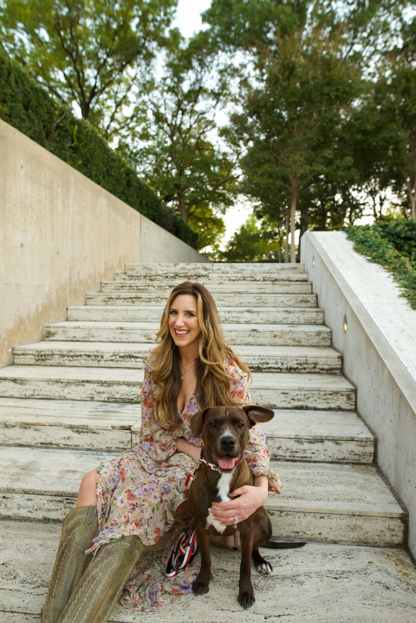 Woman sitting on stairs with a dog, outdoors, trees in background, smiling, daytime.