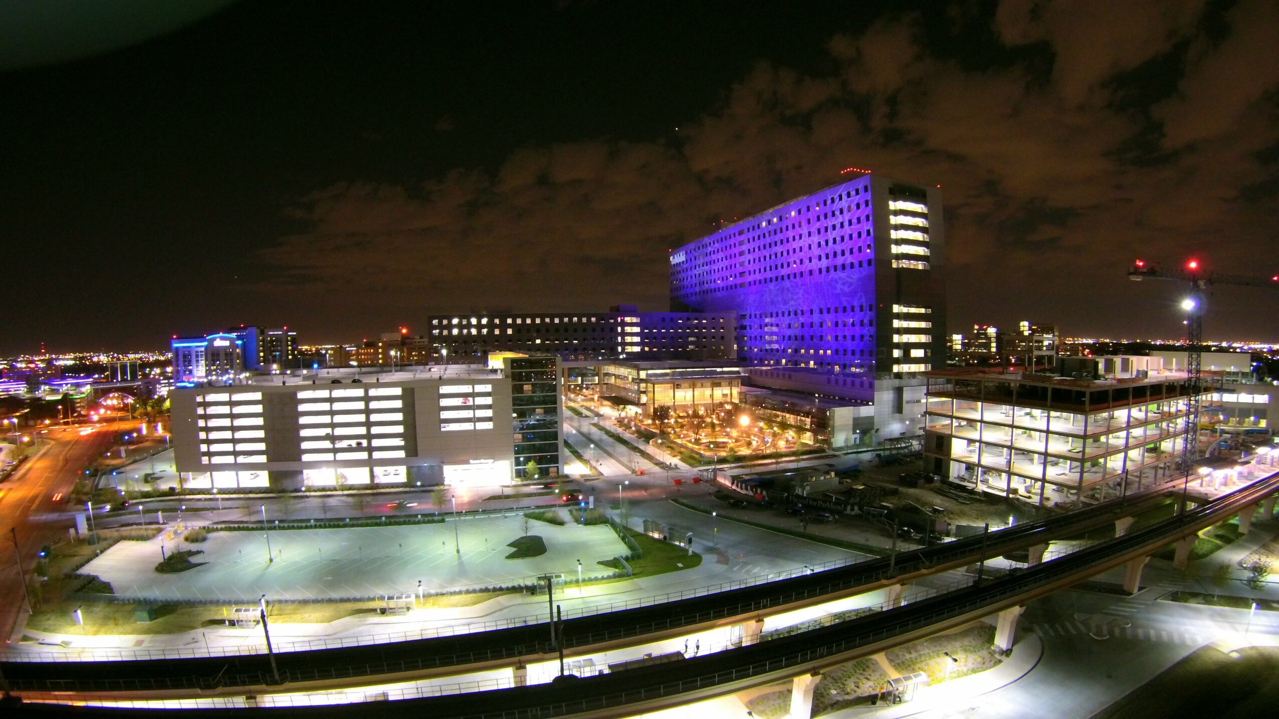 Night cityscape with illuminated buildings, a purple-lit high-rise, and a parking lot, under a cloudy sky.