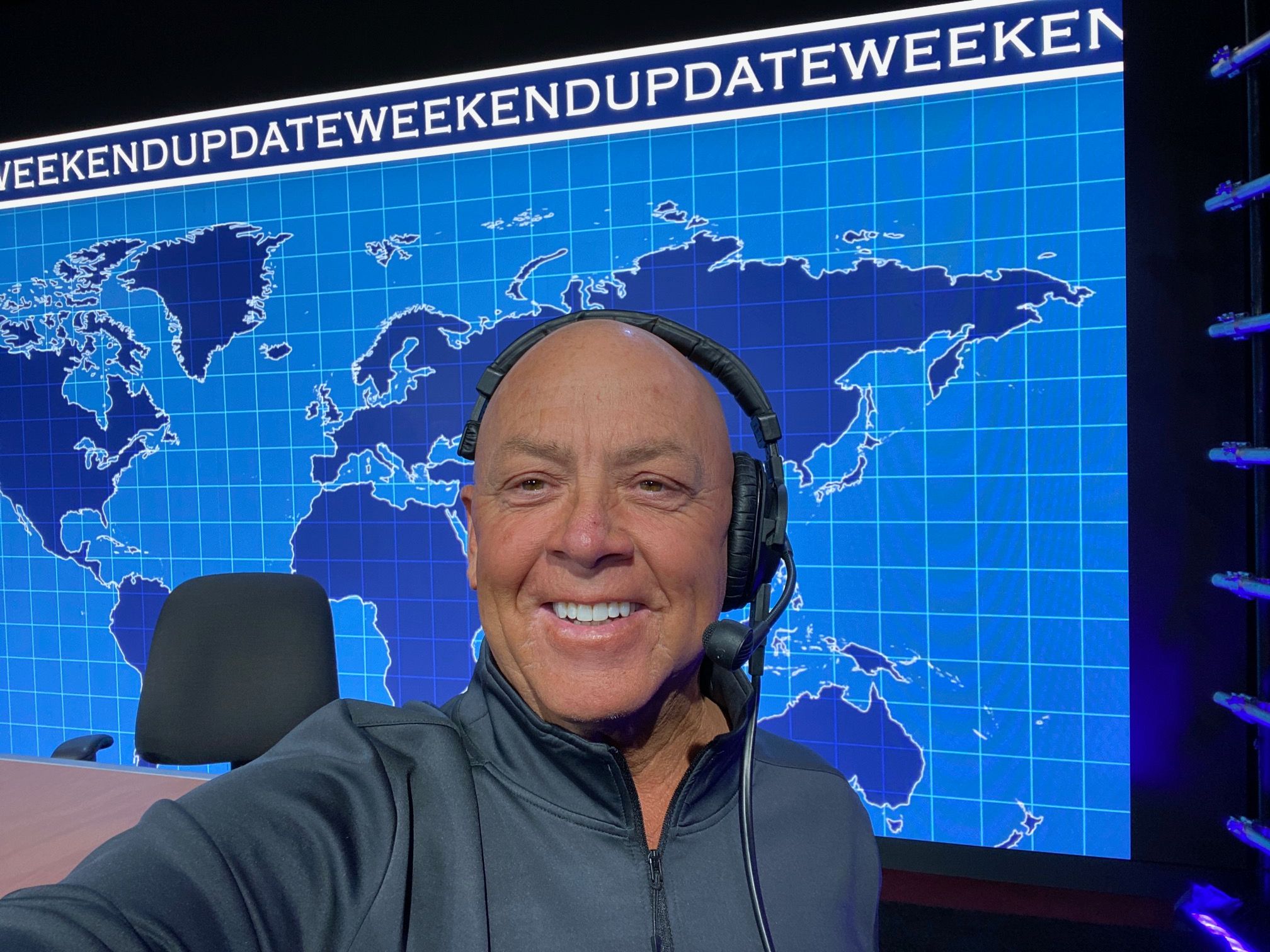 Person smiling, wearing headphones, in front of a world map display with blue background, upside down.