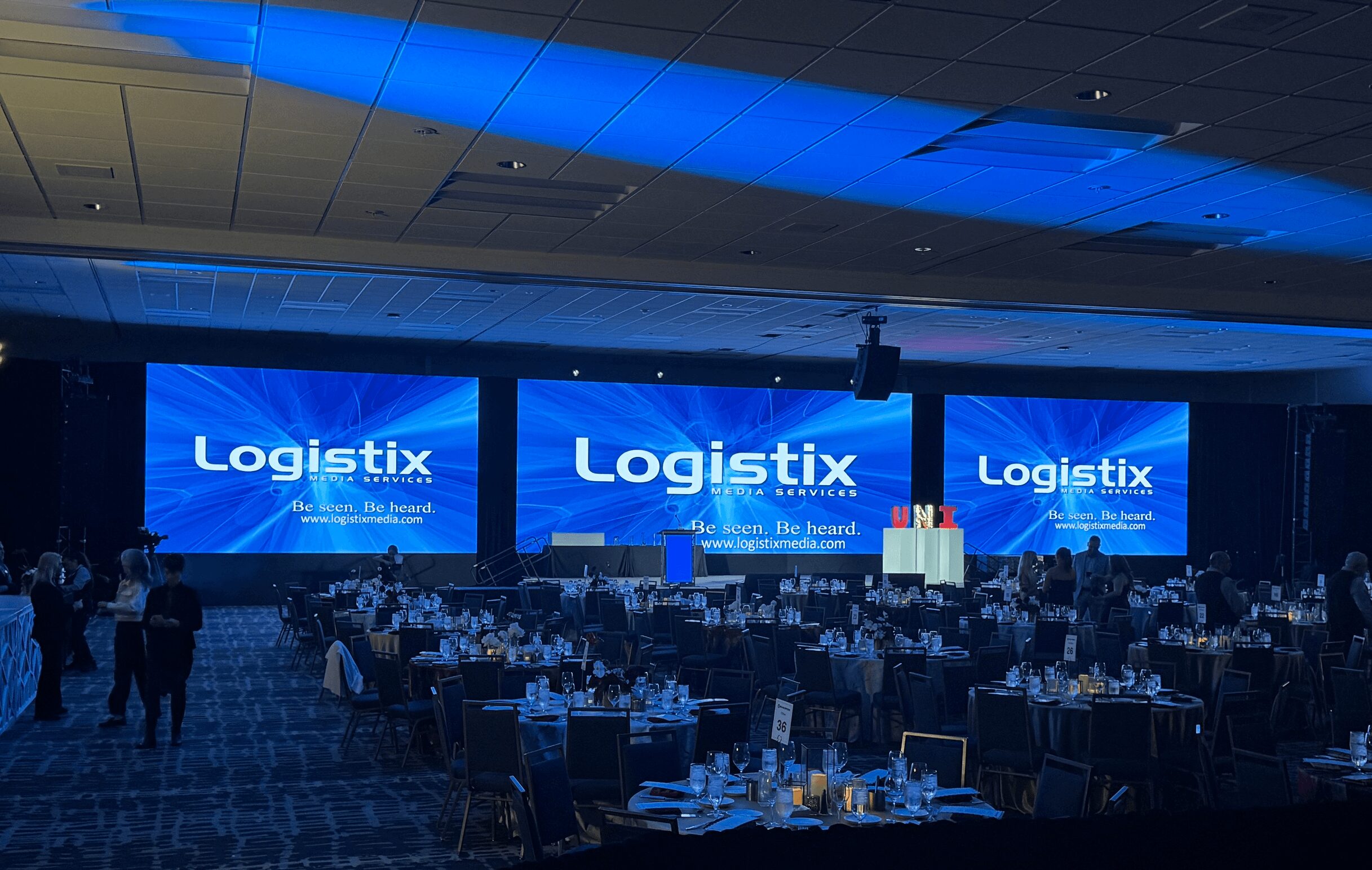 Large conference room with three screens displaying 'Logistix' and a stage with a podium, tables, and chairs.