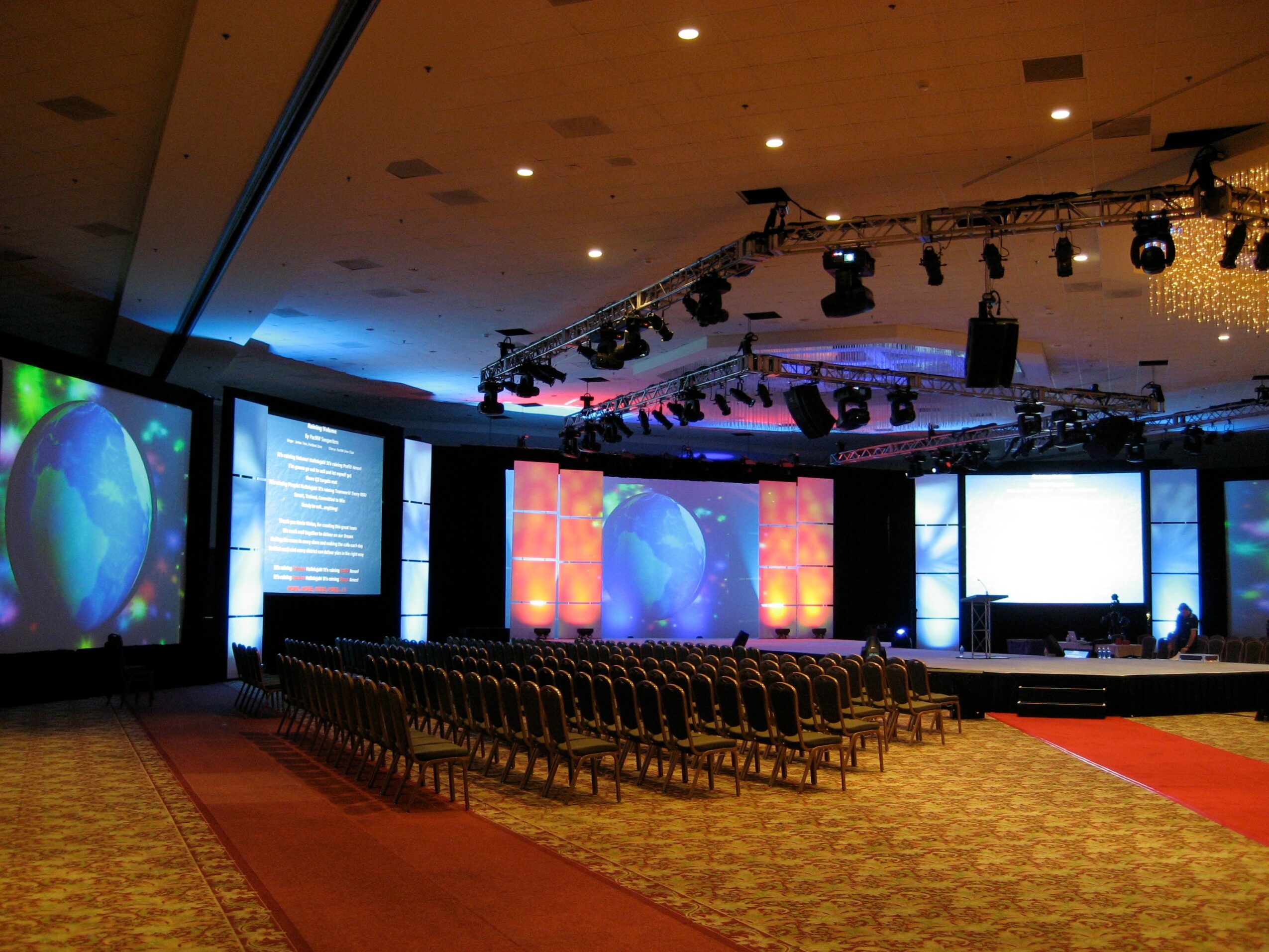 Conference room with rows of chairs facing a stage with large screens and lighting equipment.
