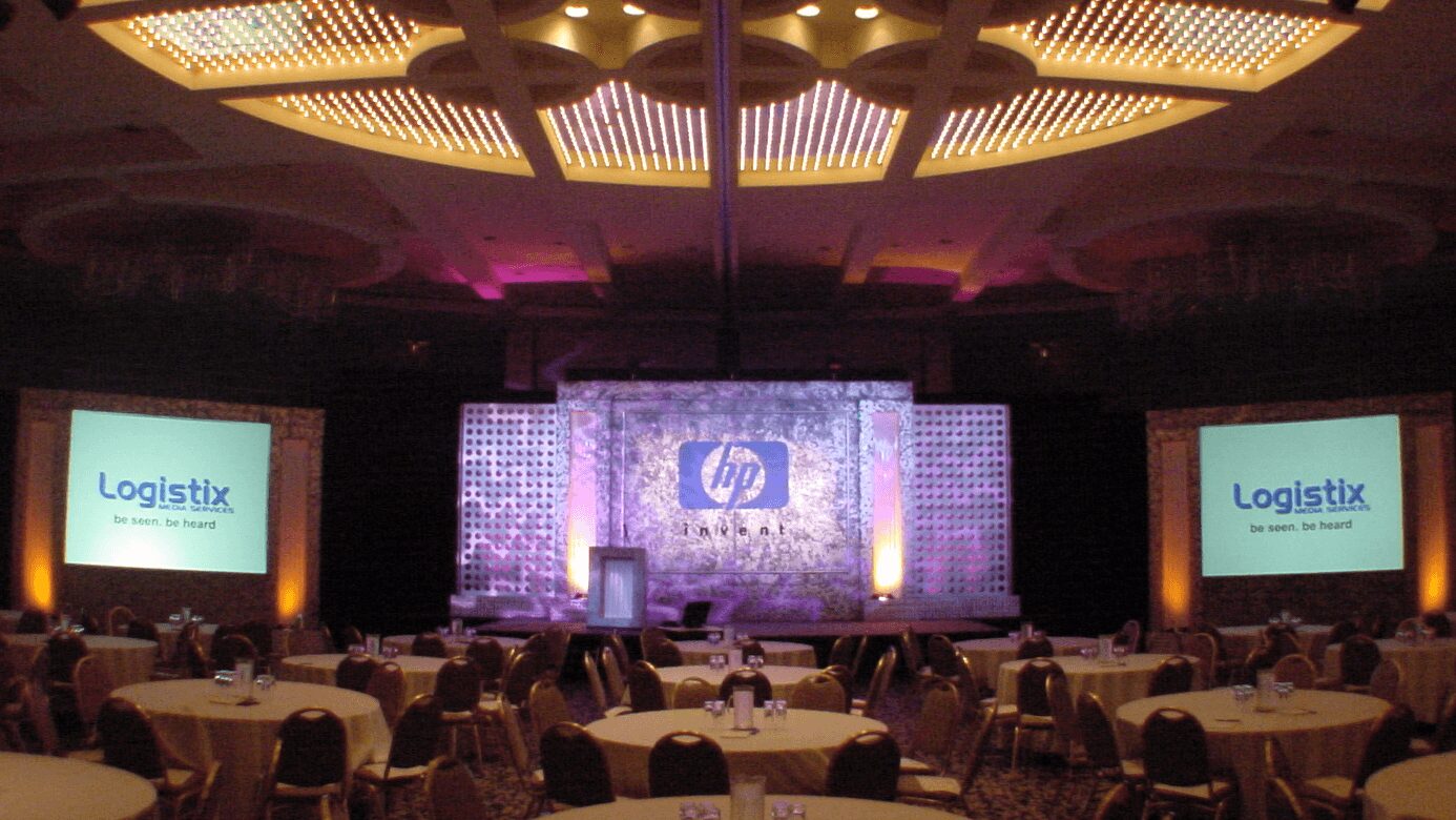 Conference room with stage, large screens displaying 'Logistix' and HP logo, audience seated, ceiling with circular lighting, dim lighting.