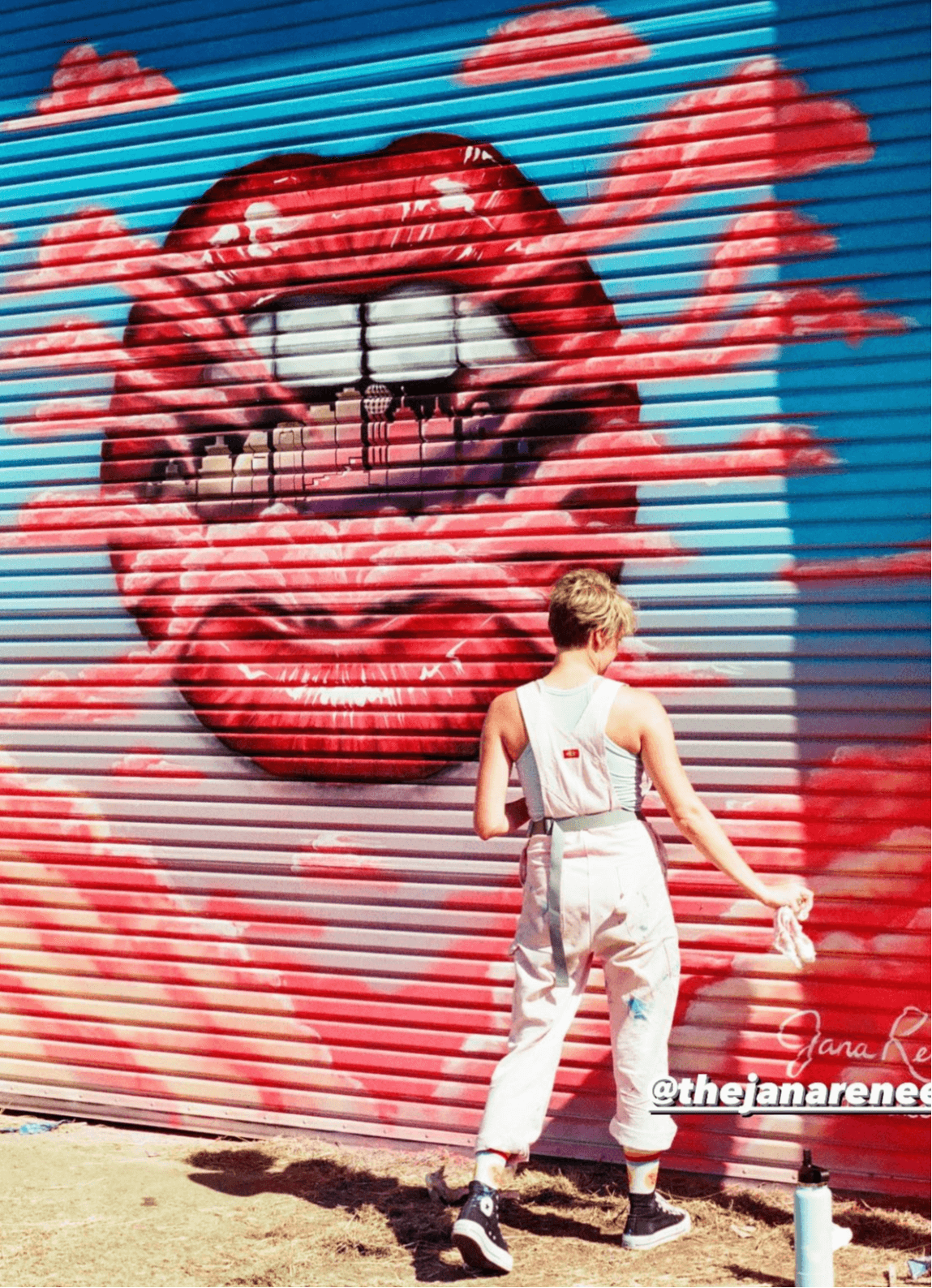 Person with short hair in white jumpsuit walking past a colorful mural of lips and clouds on a wall.