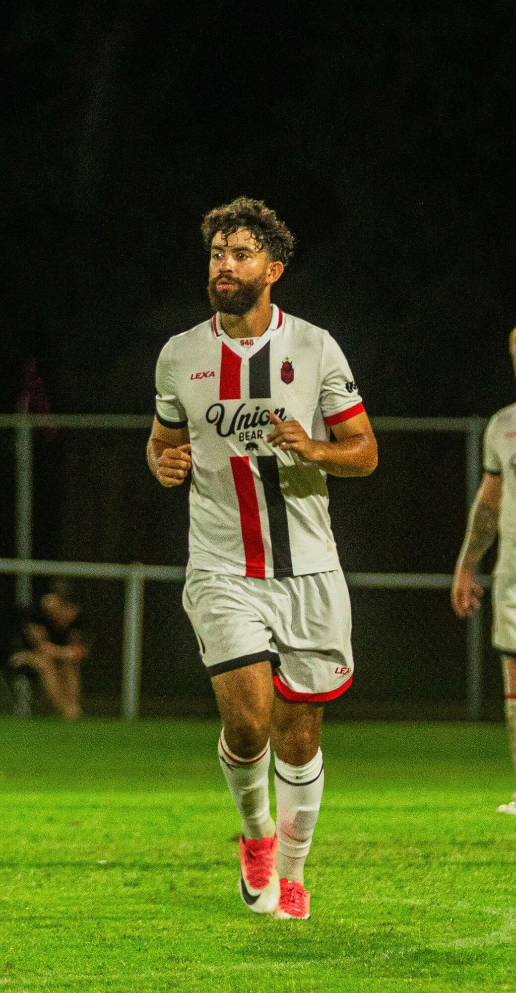 Soccer player running on field wearing white uniform with red and black stripes, pink shoes, and beard.