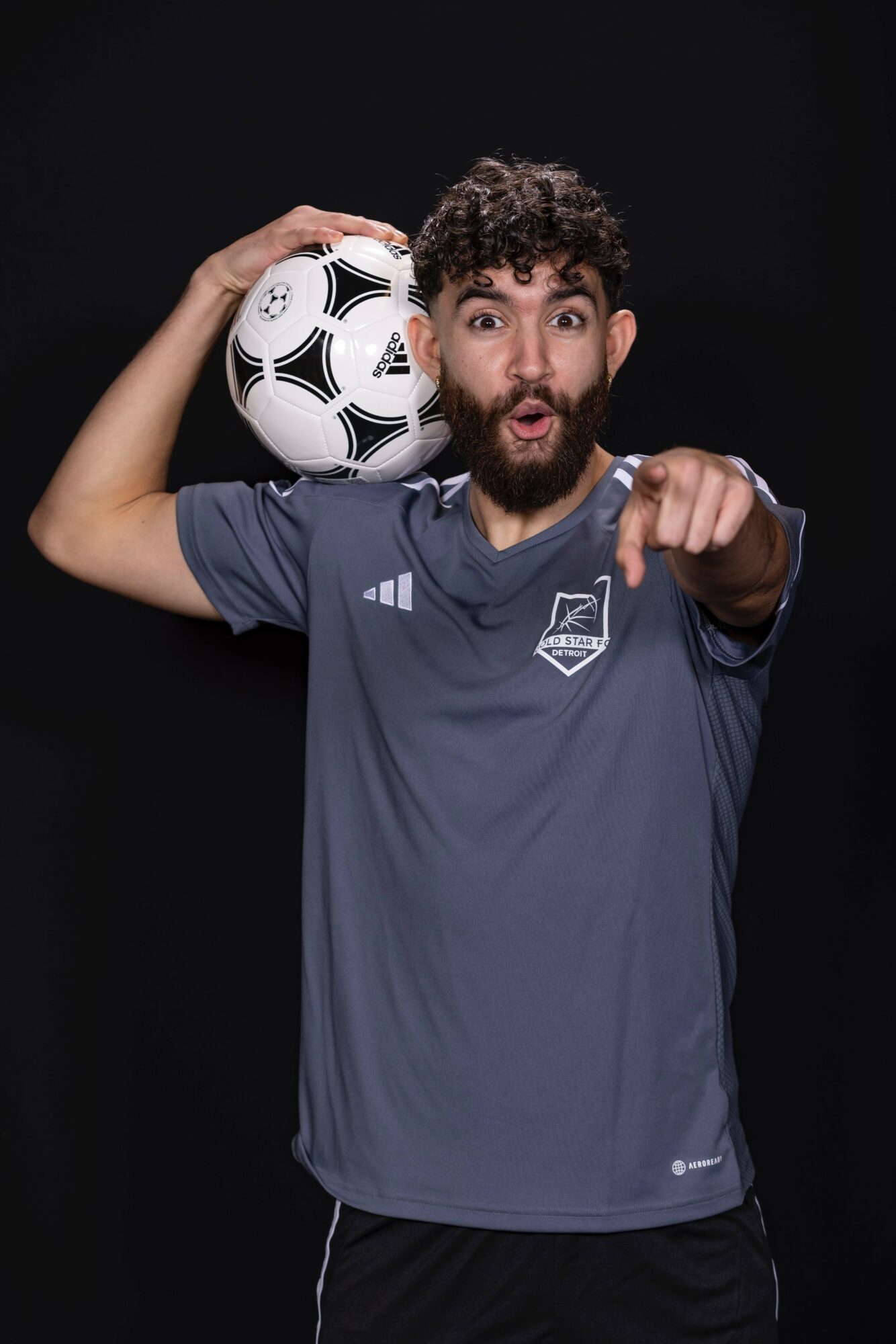 Man with curly hair and beard pointing forward, holding a soccer ball on his shoulder, wearing a dark sports jersey, black background.