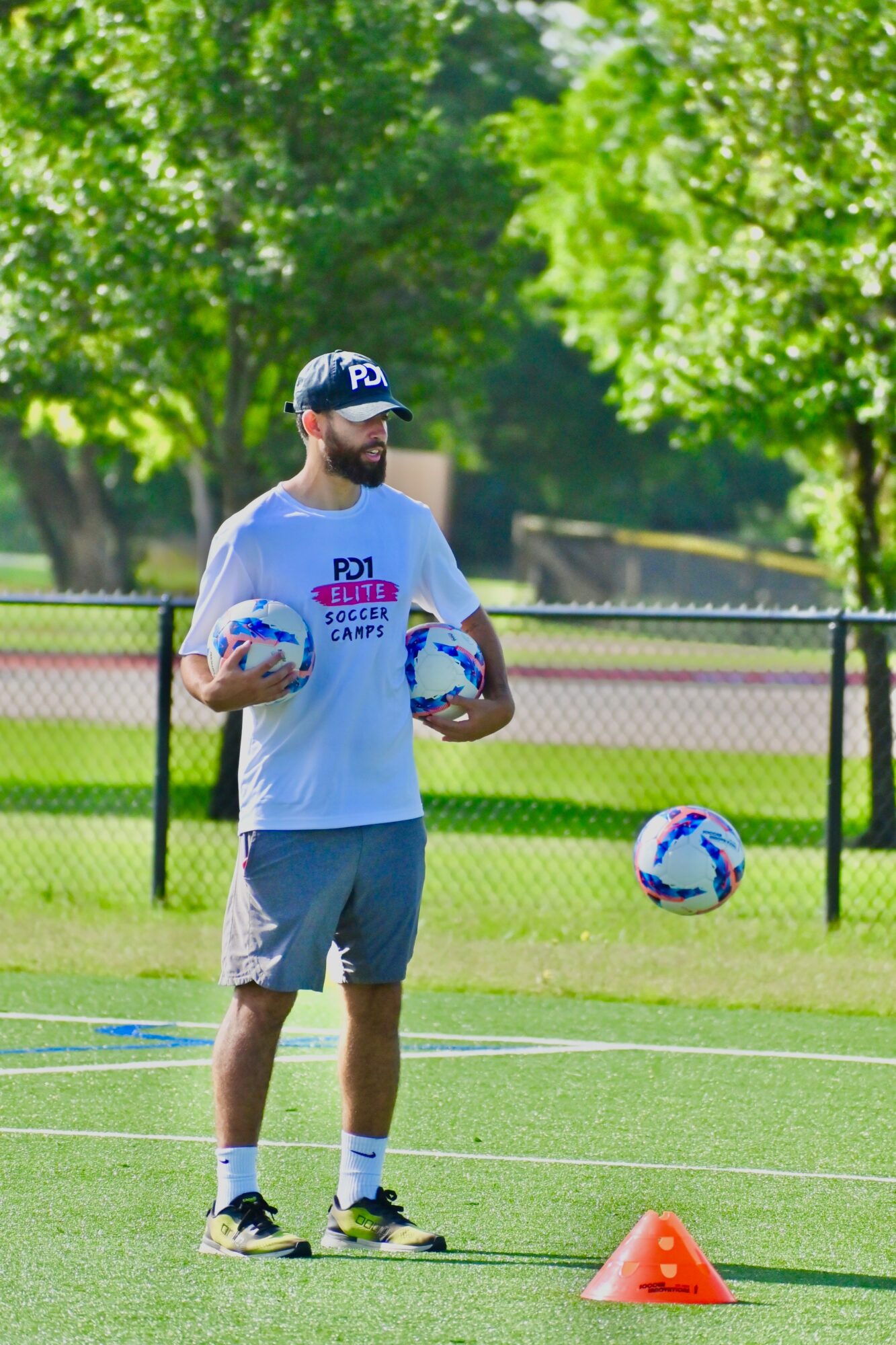 Man with a beard and sunglasses holding two soccer balls on a field with trees and a fence in the background.