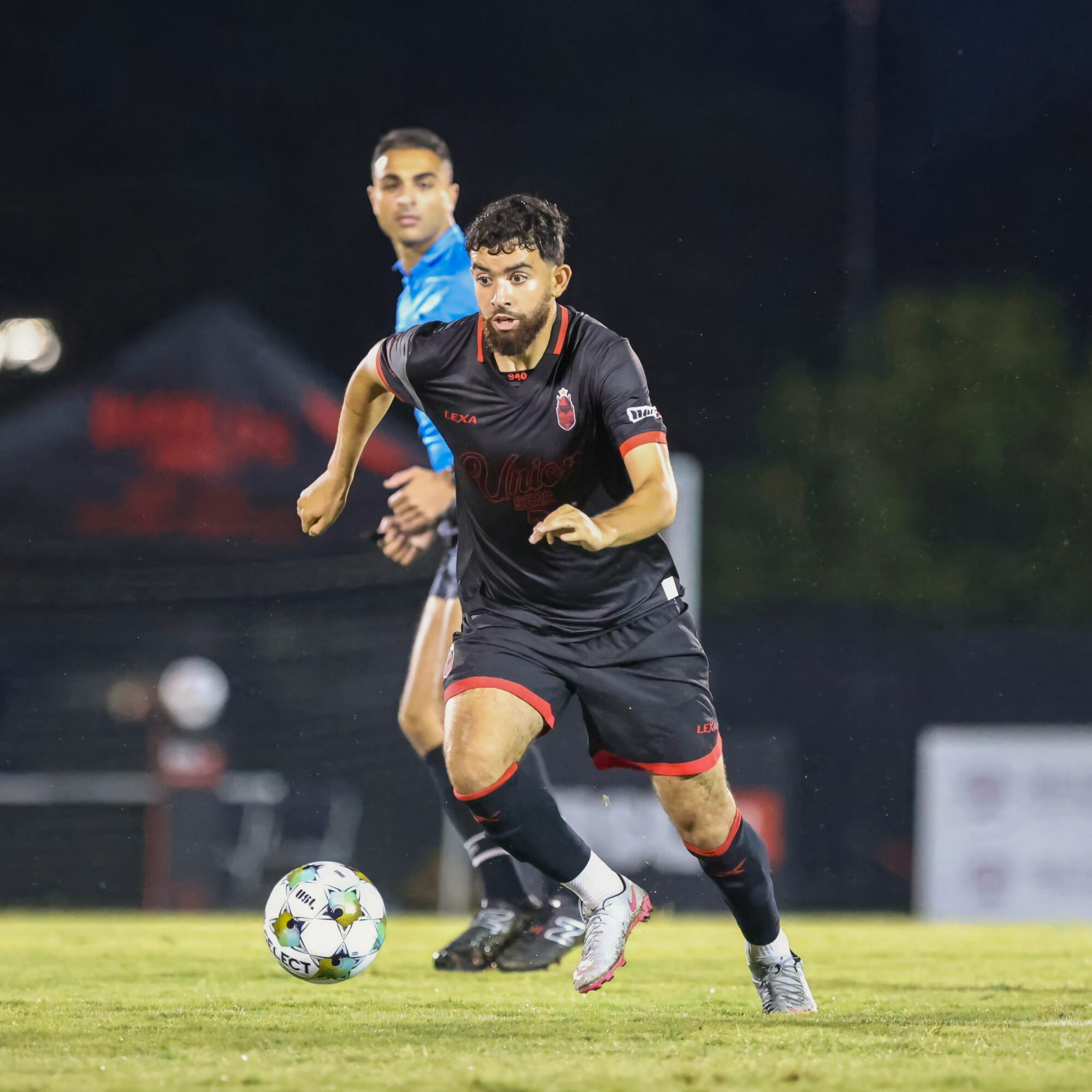 Soccer player in black uniform runs with ball on field during night game, with another player in background.