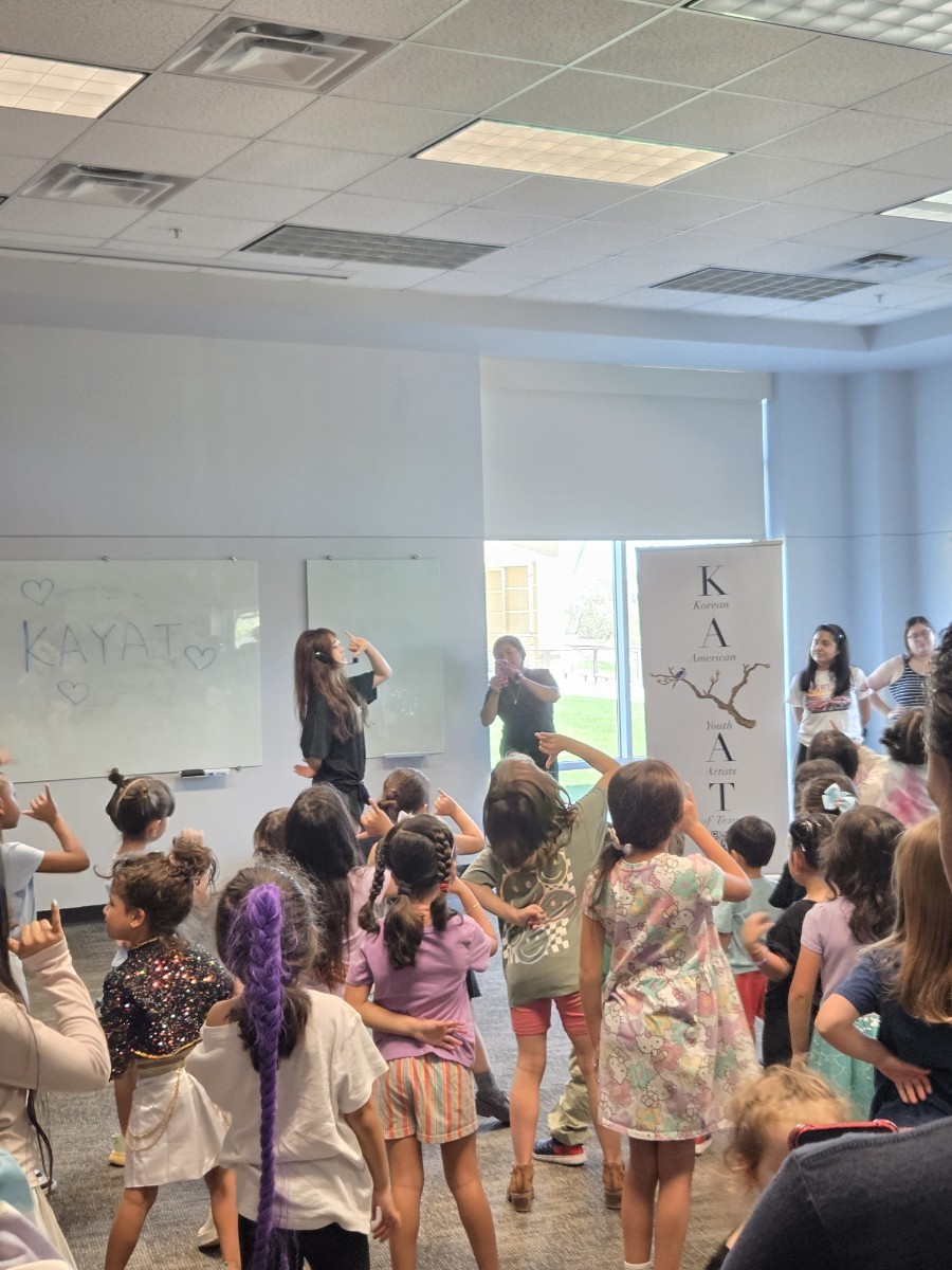 Group of children and adults in a classroom, some standing and some sitting, with a whiteboard and windows in the background.