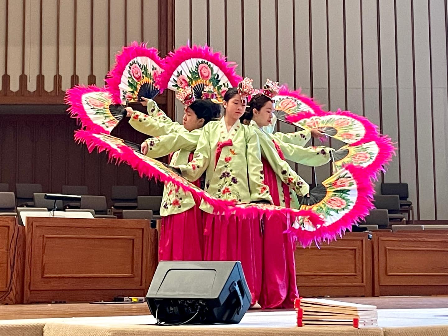 Four women in traditional clothing perform a dance with large, colorful fan-shaped props on stage.