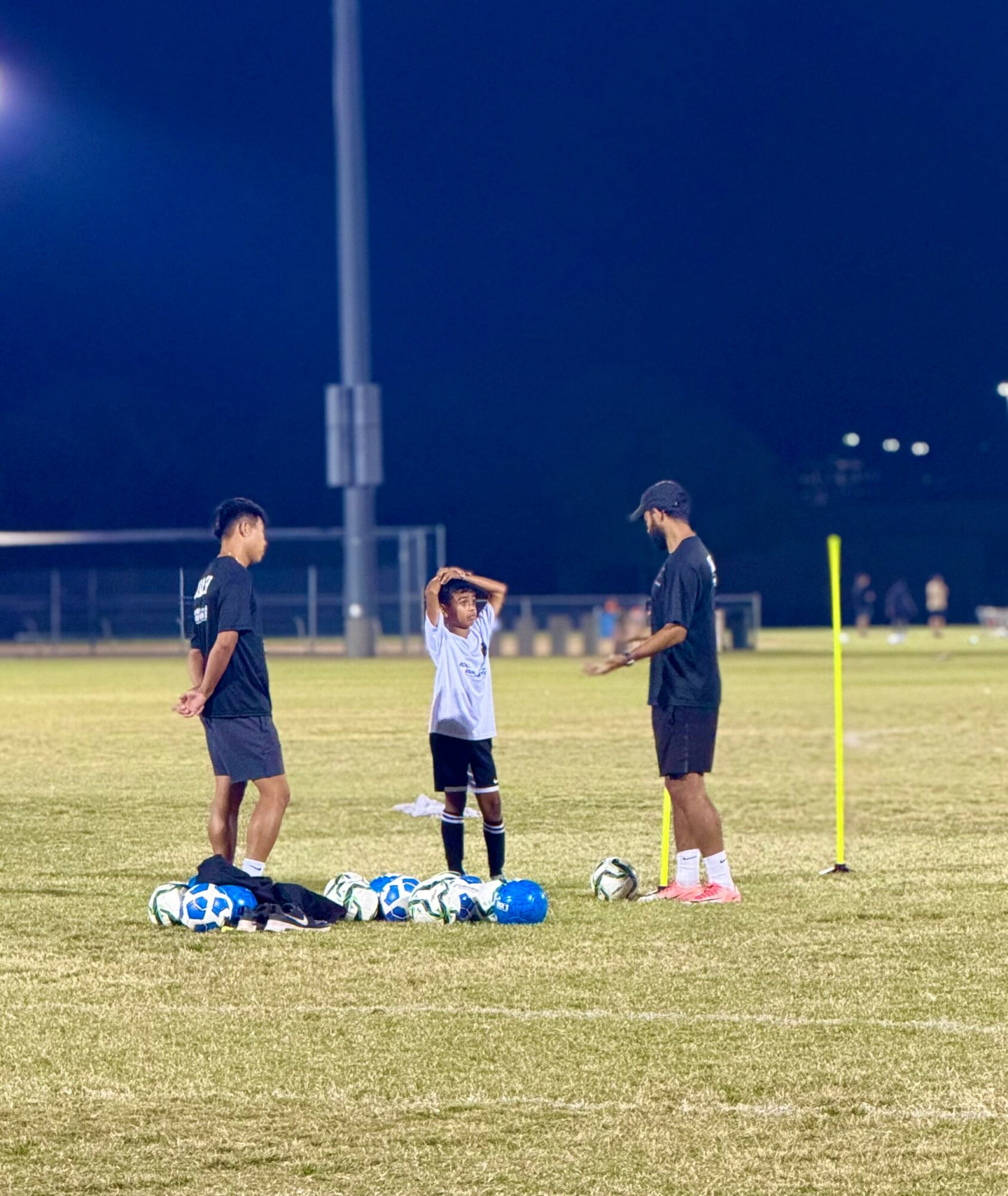 Three people, two adults and a child, standing on a soccer field at night, with soccer balls and equipment on the ground.