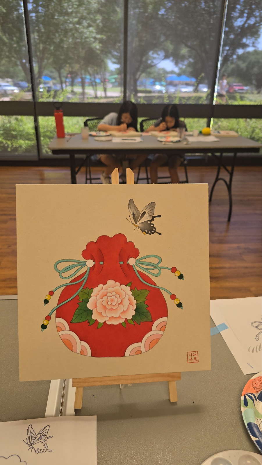 Two children sit at a table by large windows, working on art projects with supplies visible. A colorful card with a floral design and a bee is in the foreground.