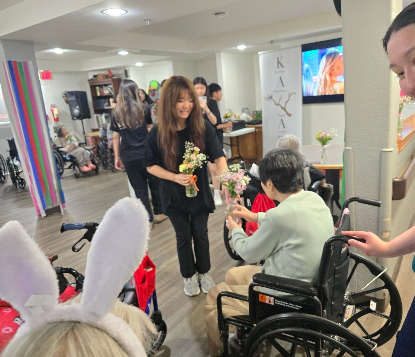 A woman with a flower bouquet interacts with an elderly person in a wheelchair, surrounded by people in a room with decorations.