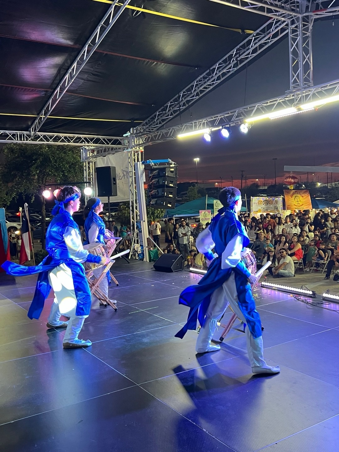 Performers in traditional costumes dance on stage under a canopy, with an audience watching at sunset.