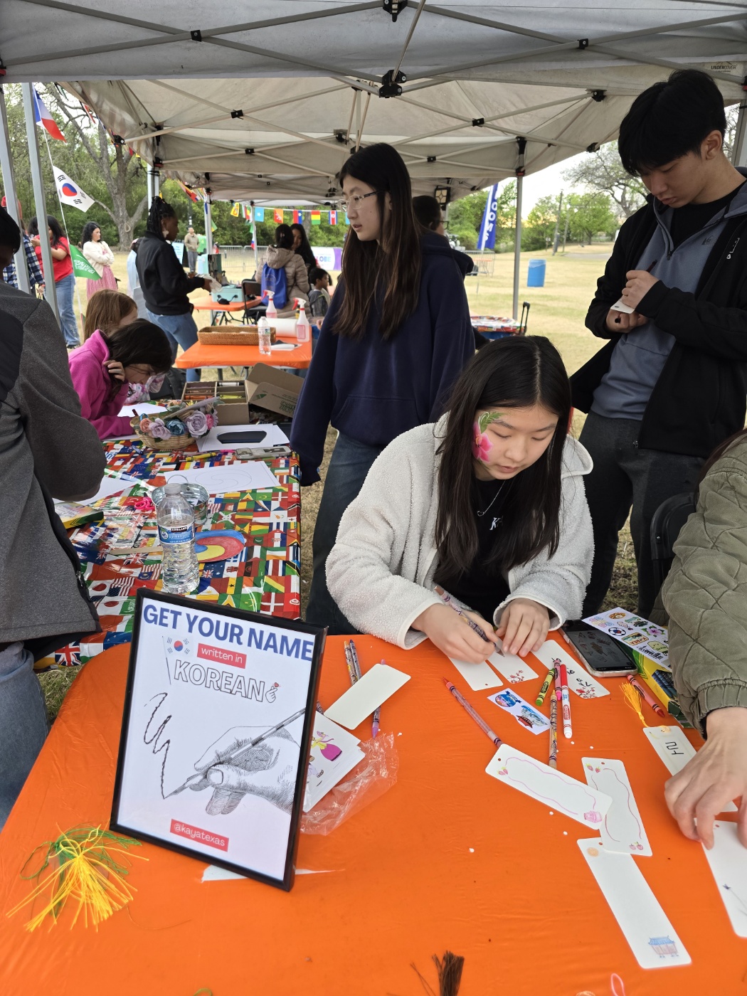 People at a booth under a canopy, with a woman writing on paper, colorful items on the table, and others browsing.