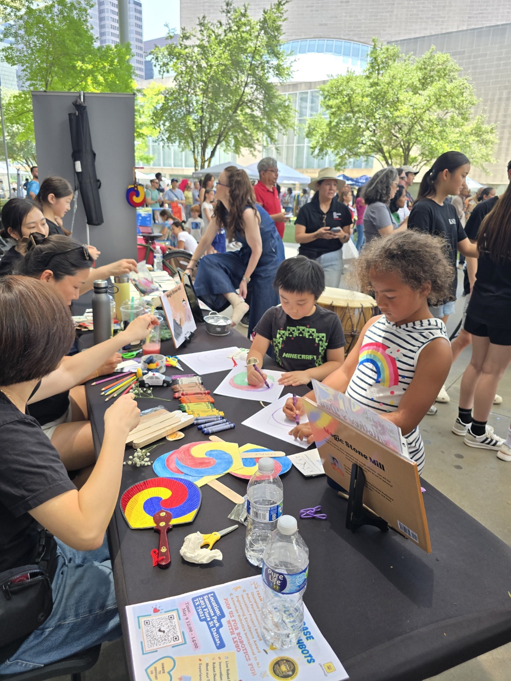Children and adults participate in a craft activity at an outdoor event, with tables, supplies, and trees in the background.