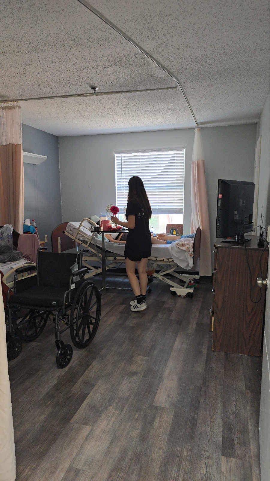 Person standing in a room with a bed, window, and wheelchair, facing the window, with furniture and a TV nearby.