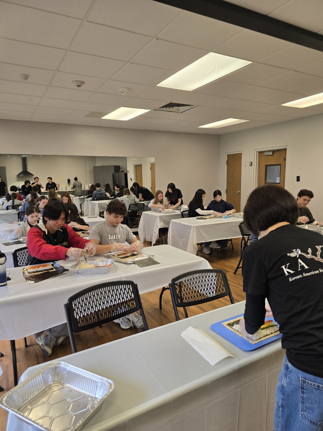 People seated at tables in a room, some working on projects, with a person standing at a table in the foreground.