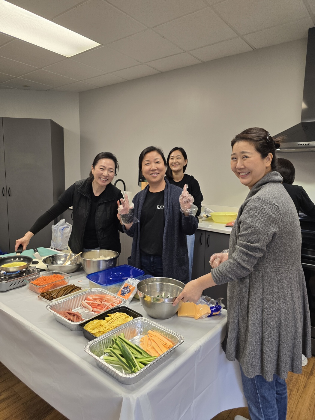 Four women in a kitchen preparing food, smiling, with various ingredients on a table.
