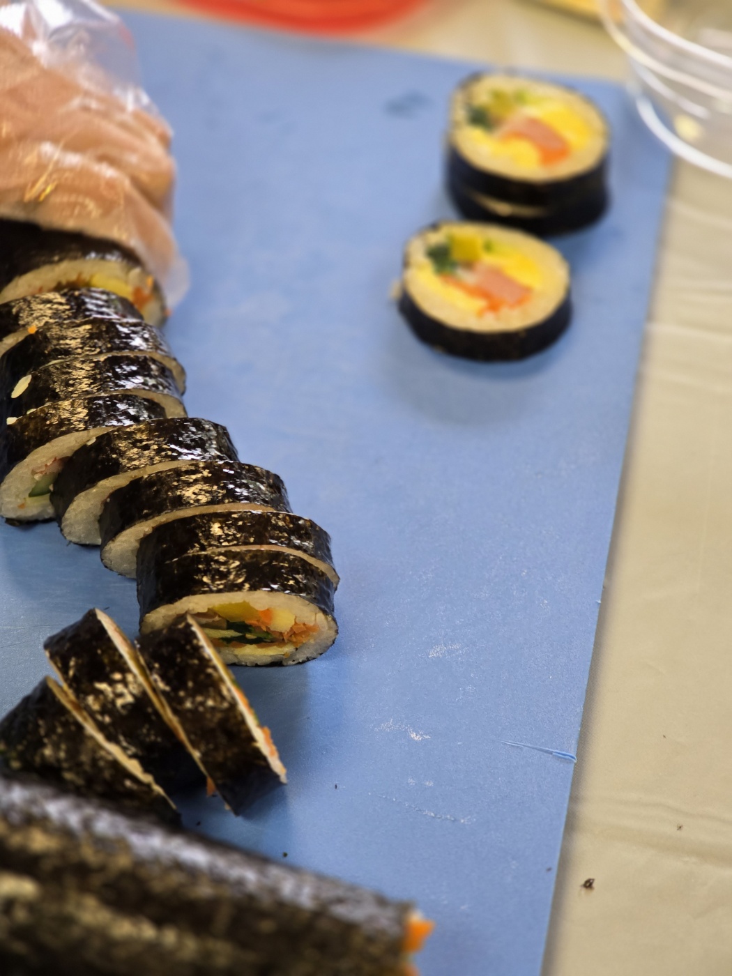 Sushi rolls on a blue cutting board with two pieces separated, colorful filling visible, and a hand in a glove nearby.