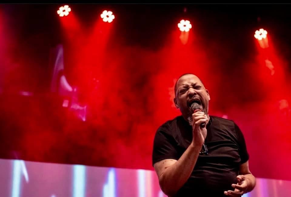 Man singing passionately on stage with red lighting and smoke effects, holding a microphone, wearing a black shirt.