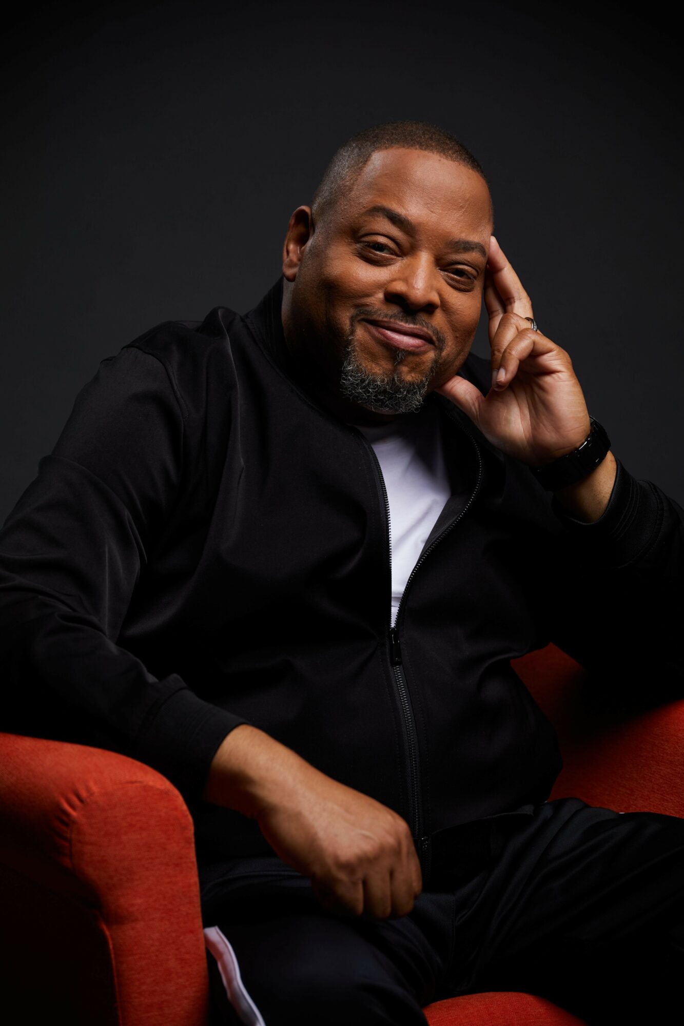 Man with a beard smiling, resting his head on his hand, wearing a black jacket, seated on a red chair.