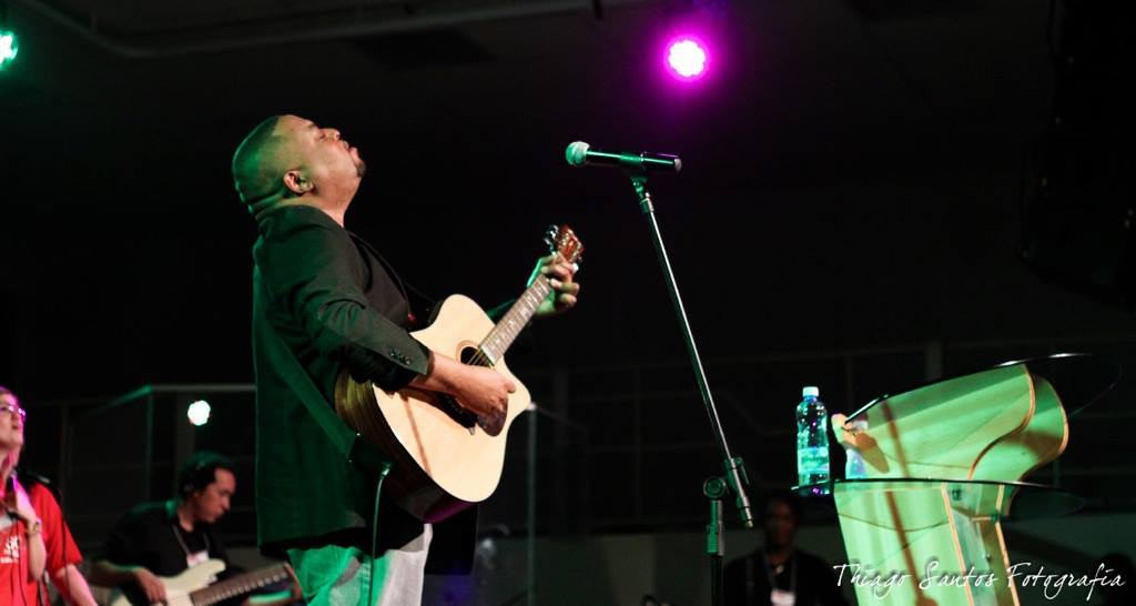 Person playing guitar on stage with microphone, purple stage lighting, and other musicians in background.