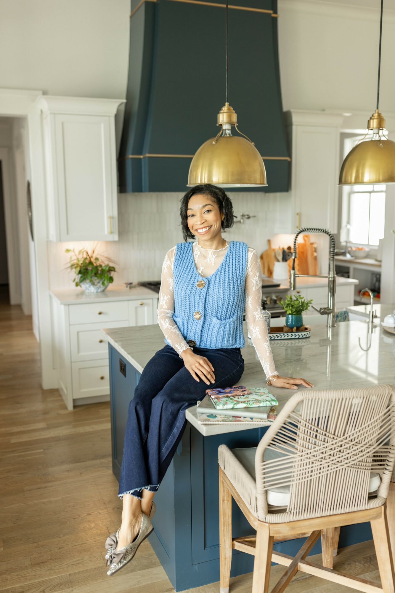 Woman sitting on kitchen island, smiling, with modern kitchen background, white cabinets, and gold pendant lights.