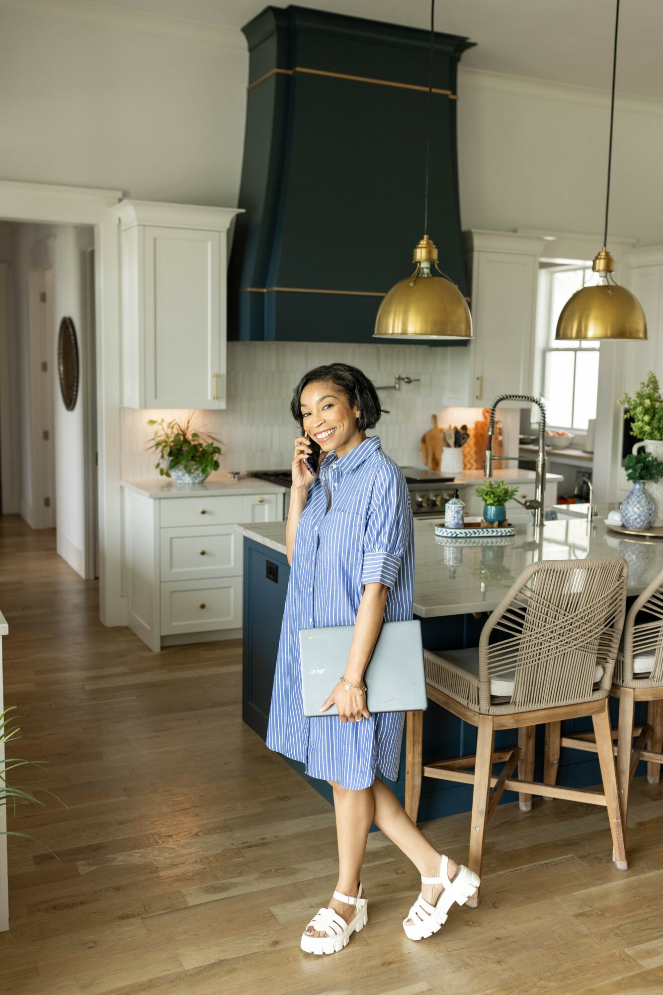 Woman in blue dress holding a laptop, standing in a modern kitchen with white cabinets and gold pendant lights.
