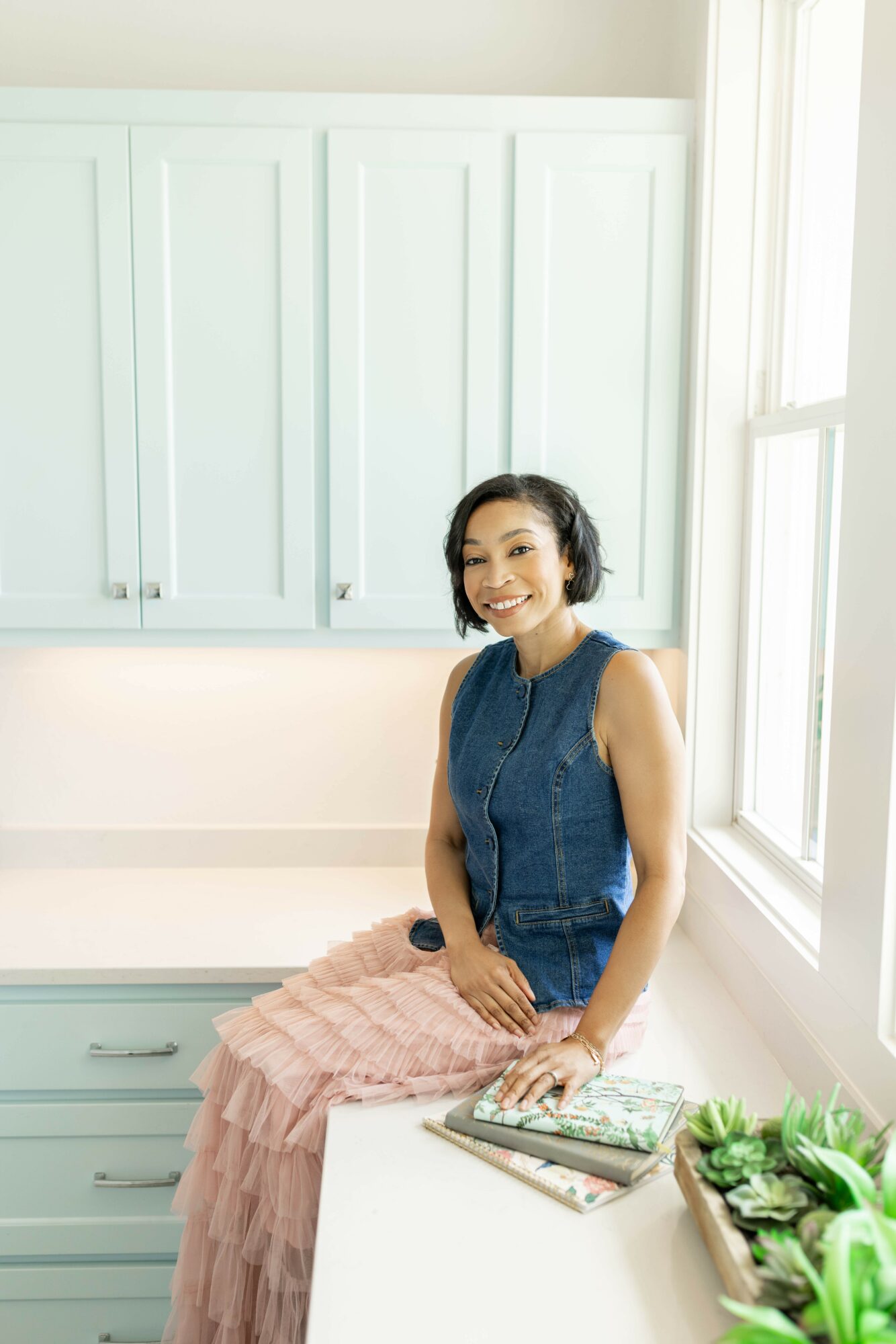 Woman sitting on a white surface near a window, smiling, with light blue cabinets behind her, holding a book, plants nearby.