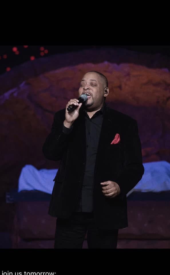 Man singing into a microphone on stage with a colorful background, wearing a black suit and red pocket square.