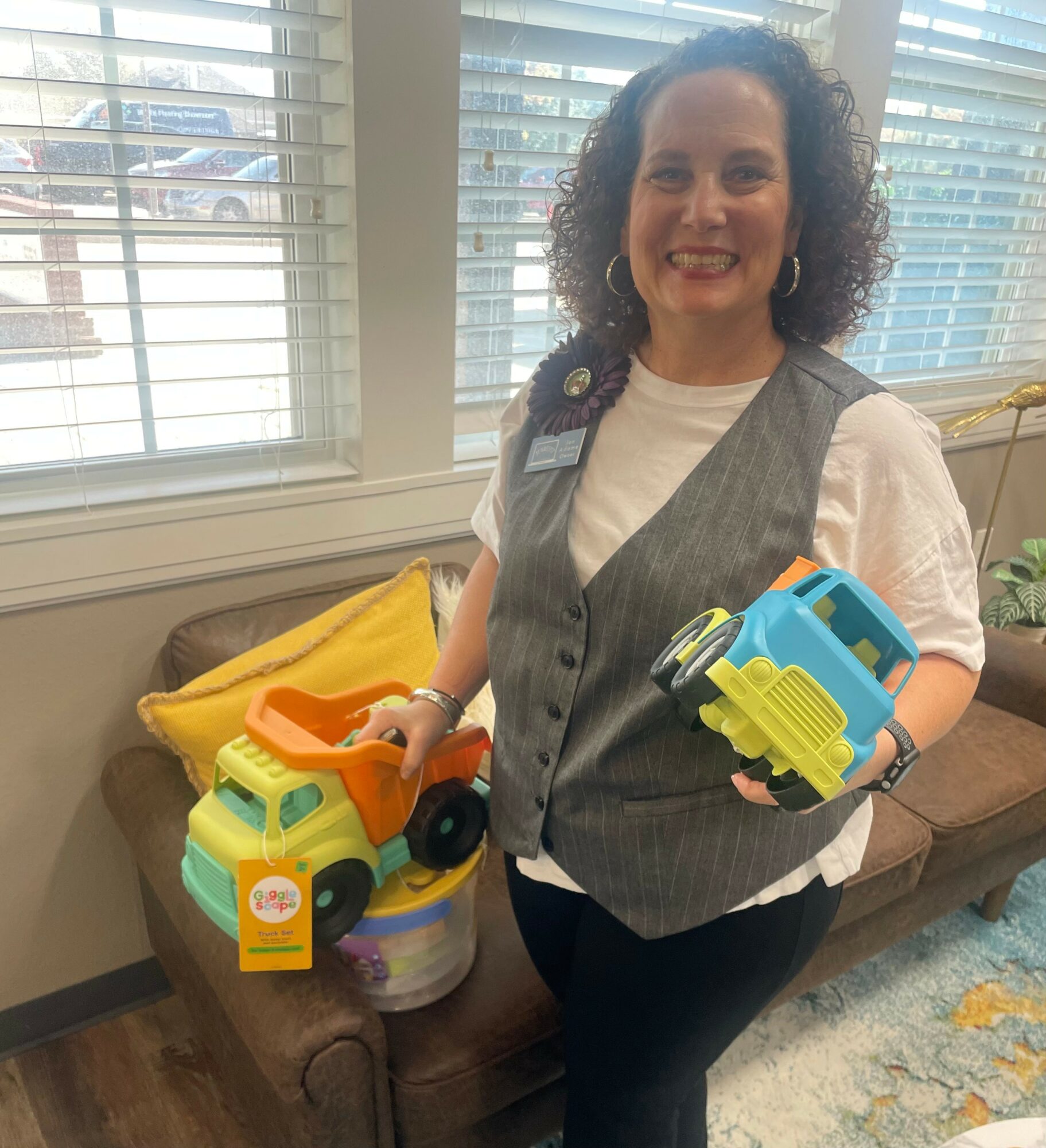 Woman with curly hair holding colorful toy trucks, standing indoors near a window with blinds, smiling.