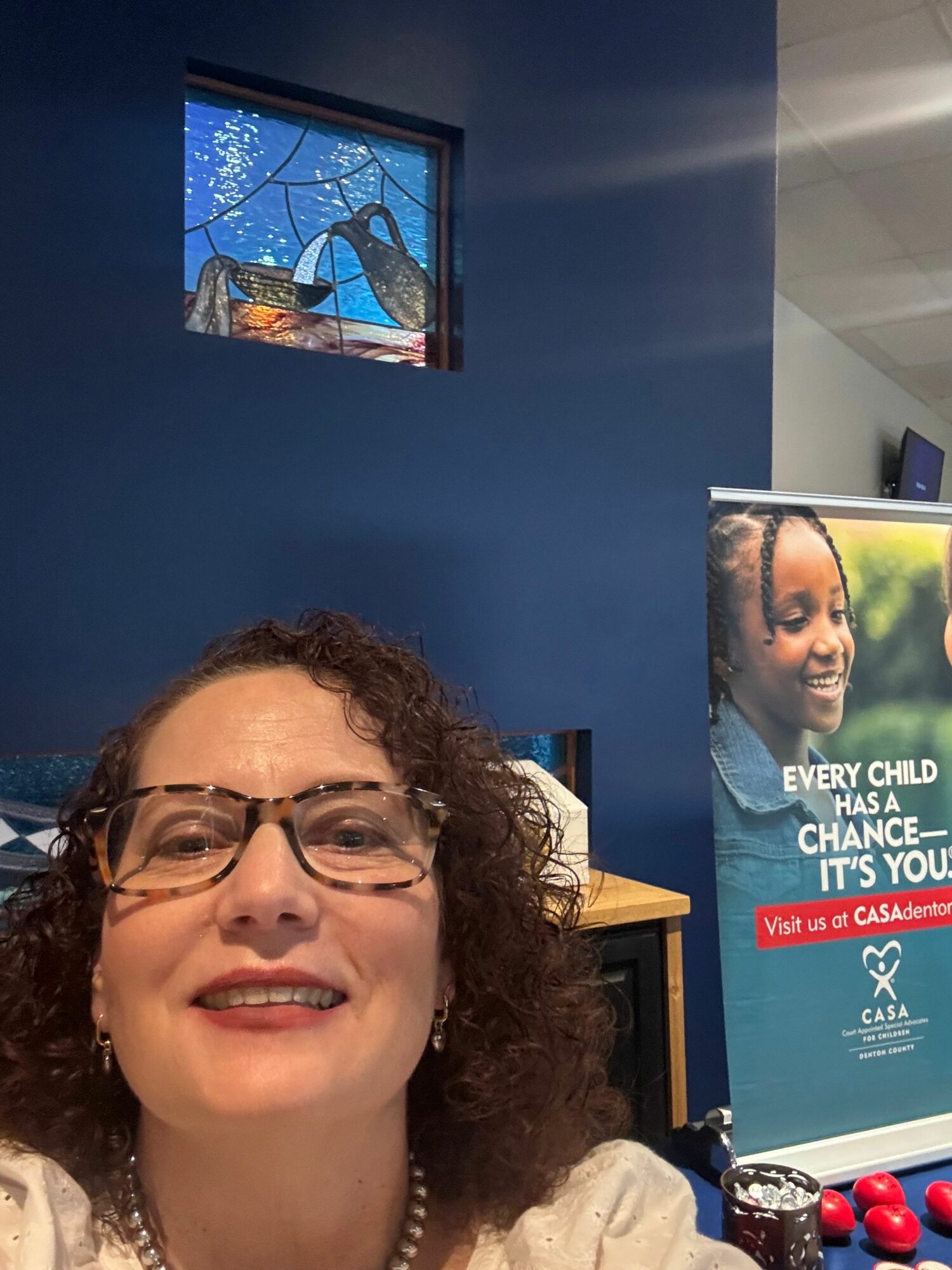 Smiling woman with glasses and curly hair in a room with blue walls and a poster behind her.