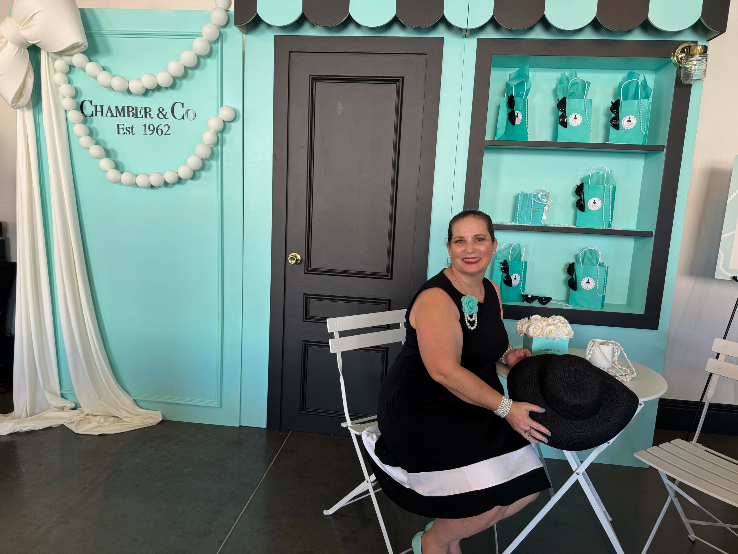 Woman sitting at a table in a teal and black room with shelves and a door, smiling, holding a black hat.
