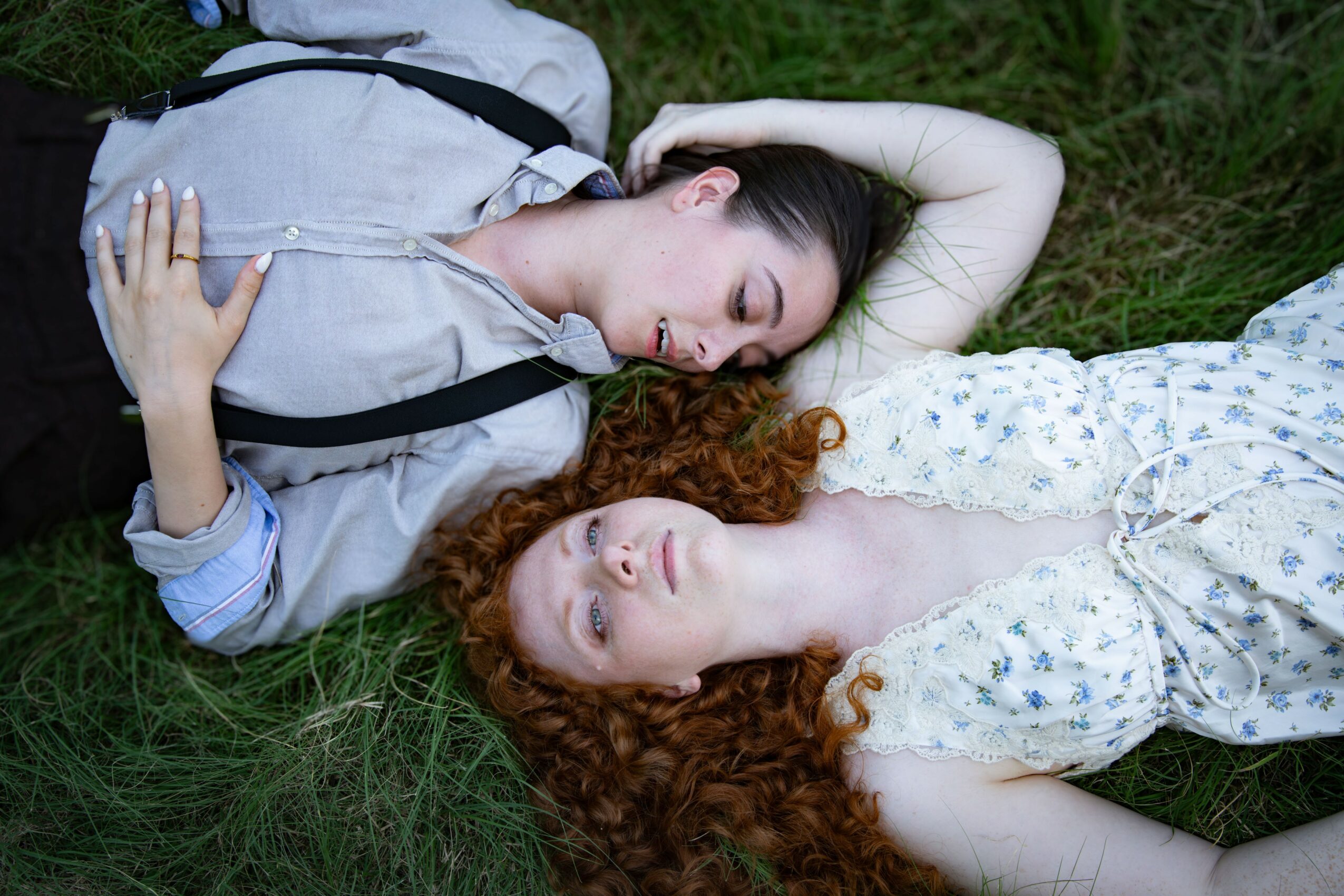 Two women with curly hair lying on grass, one with dark hair and the other with red hair, eyes closed.