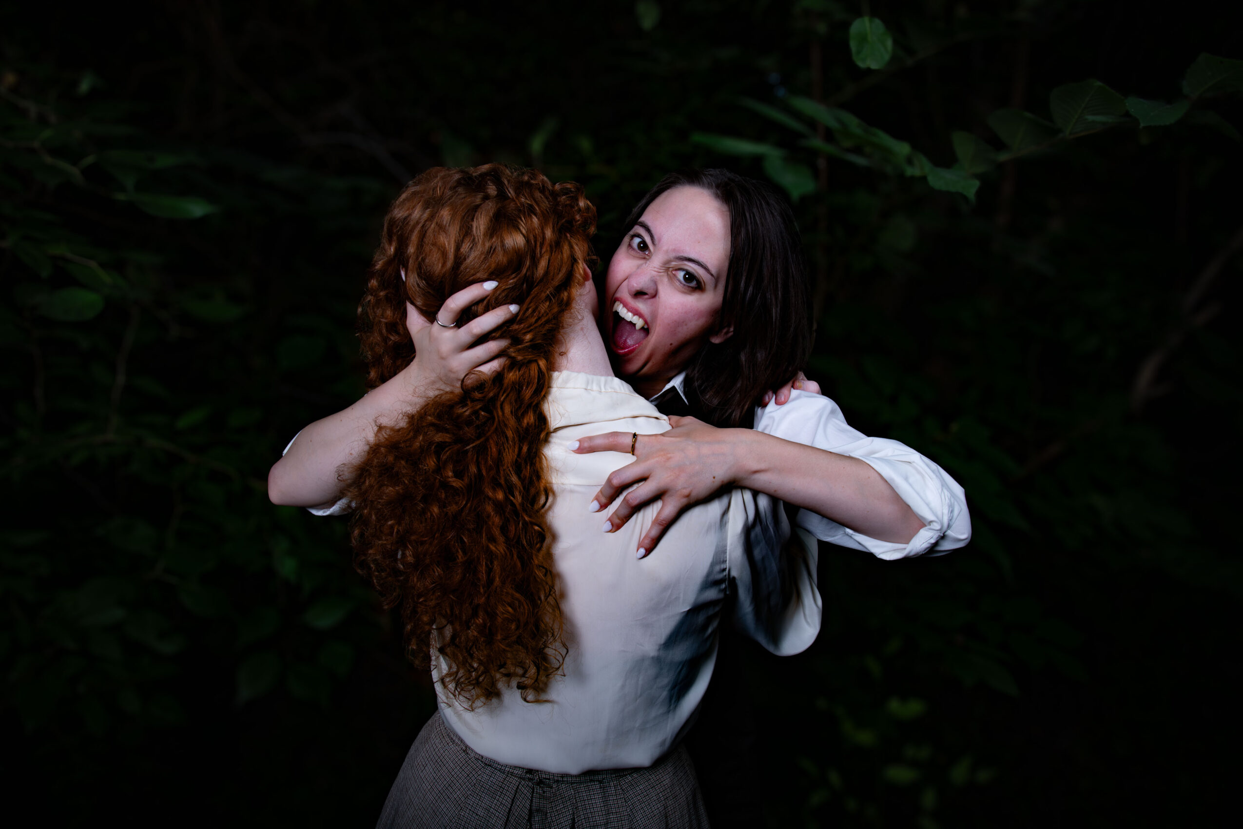 Two women hugging outdoors, one with long black hair and the other with curly red hair, smiling and making a playful face.