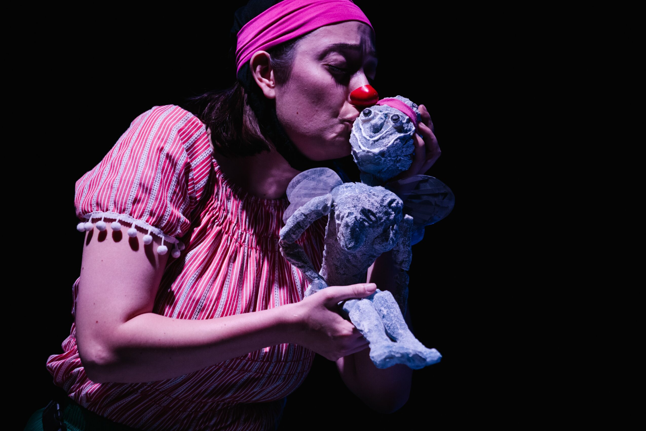 Clown with pink headband and striped dress holding a stuffed elephant and mouse, face close to a stuffed mouse, dark background.