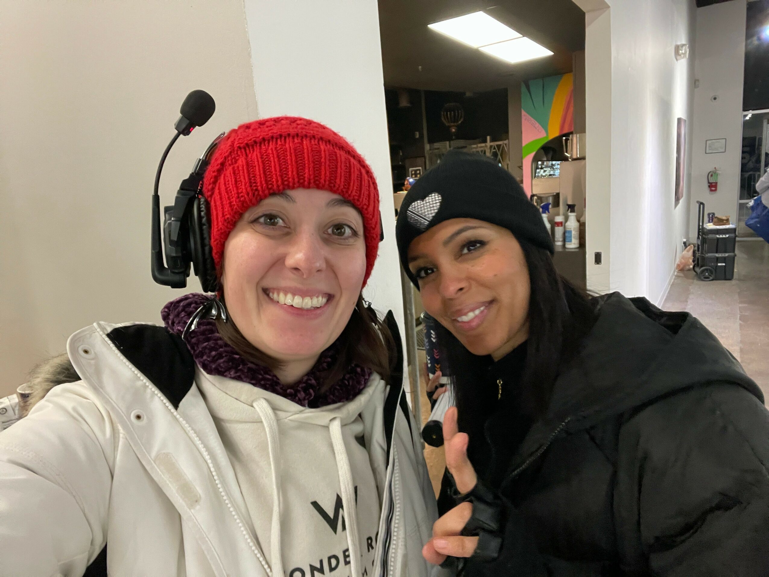 Two women smiling, one wearing a red beanie and headphones, the other in a black jacket, in an indoor setting.