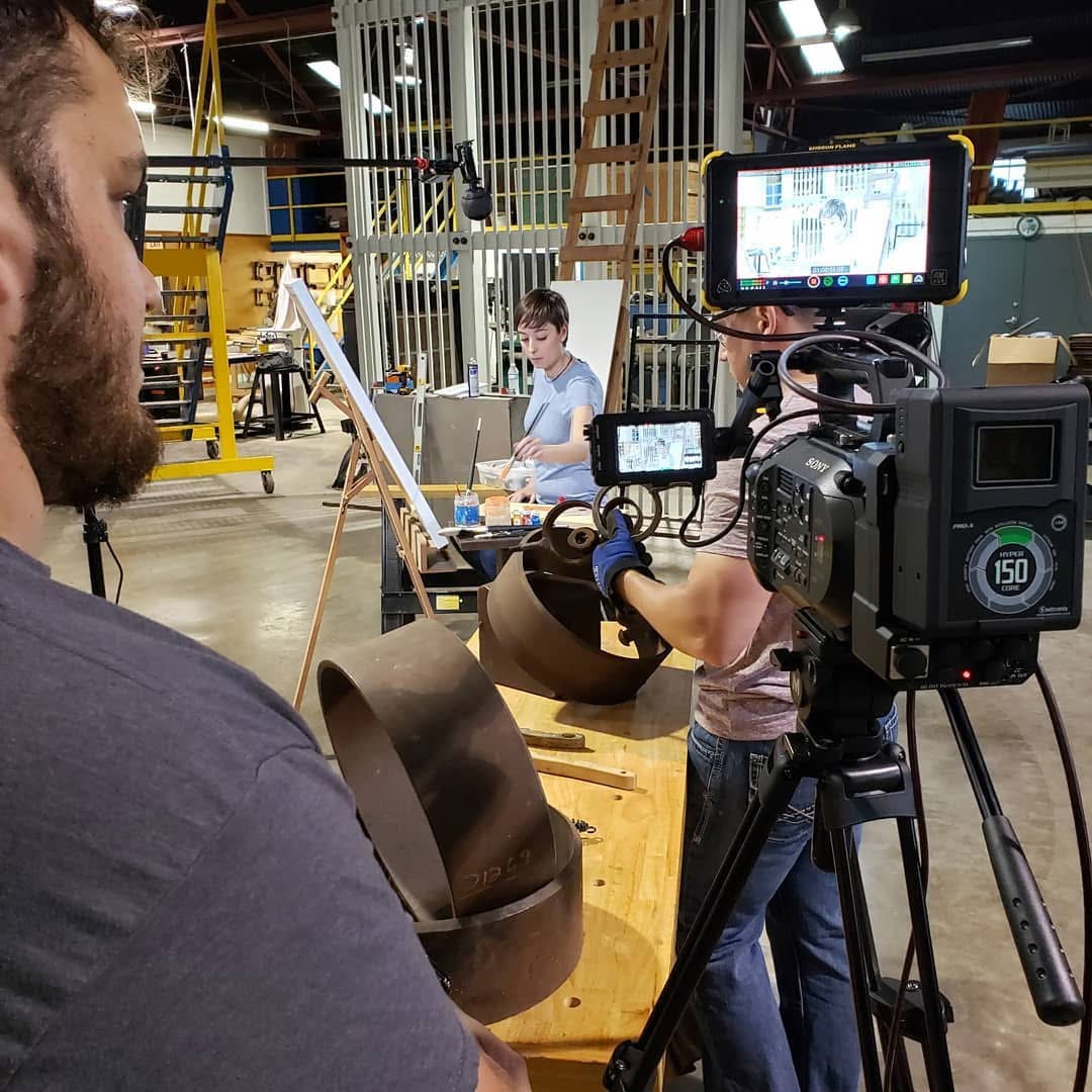 Person operating a professional video camera in a workshop with a person working on a laptop in the background.