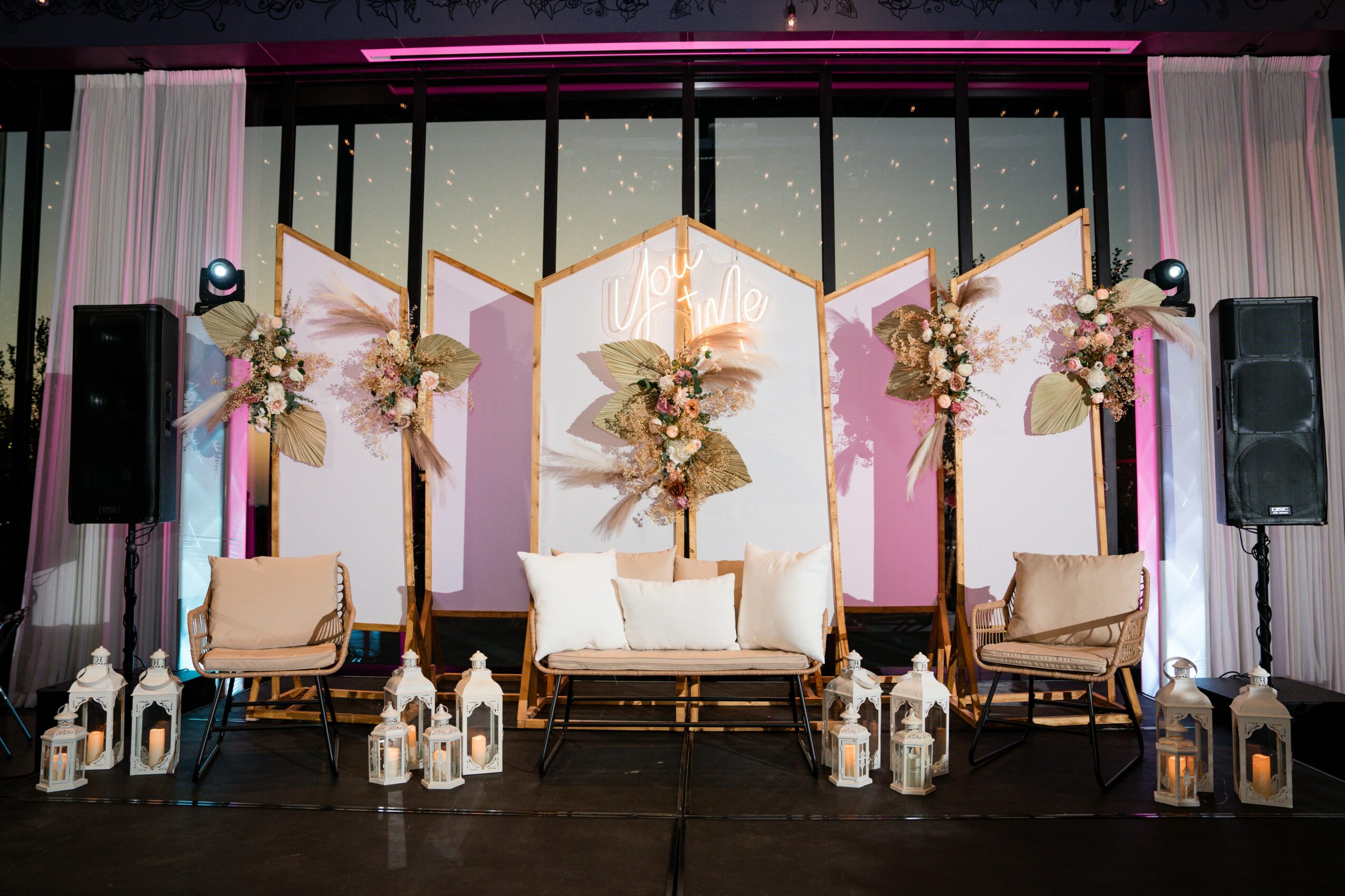 Stage with floral arrangements, white and beige furniture, lanterns, and a backdrop of large windows with curtains.