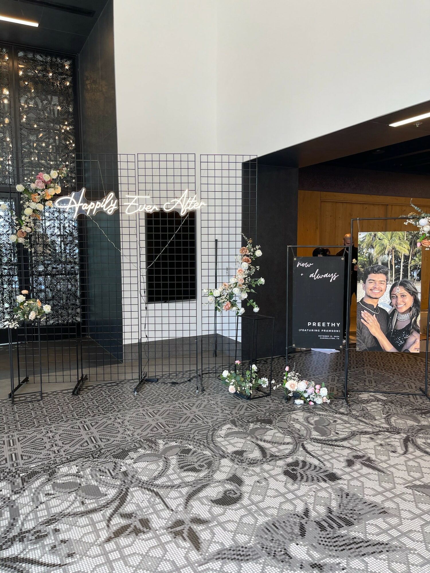 Indoor event space with floral arrangements, a black sign, and a photo display, with patterned floor and white wall.