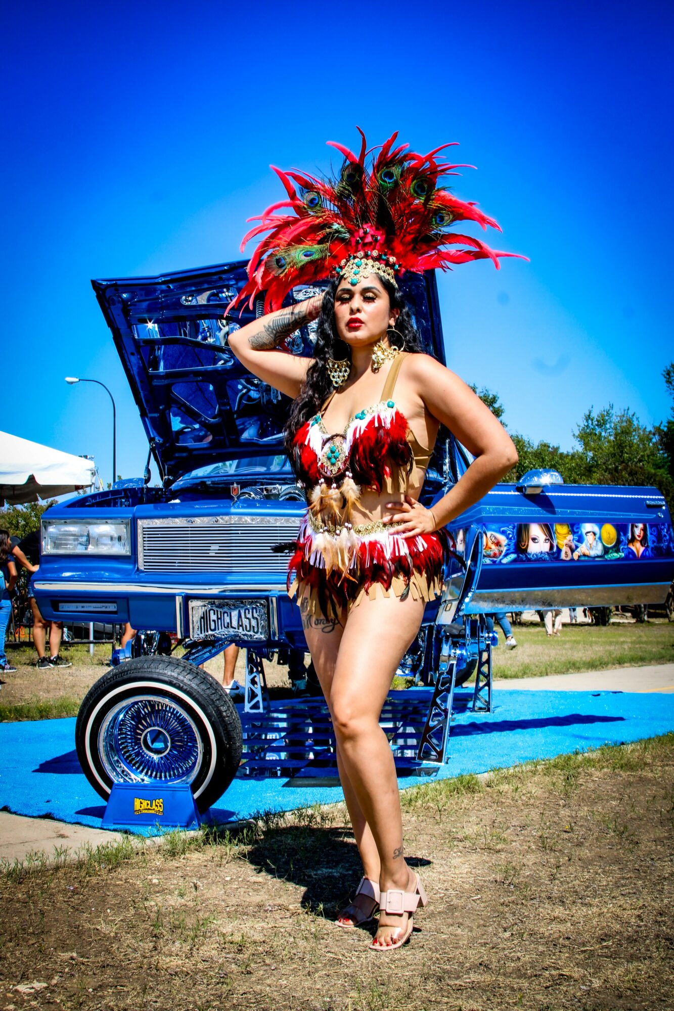 Woman in elaborate costume with feathers and beads, standing outdoors near a blue vehicle, with a clear blue sky in background.