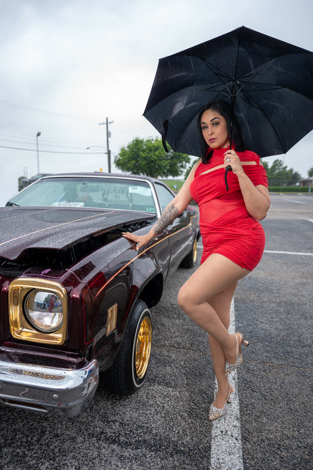 Woman in red dress holding black umbrella next to vintage car on rainy day, standing on wet parking lot, overcast sky