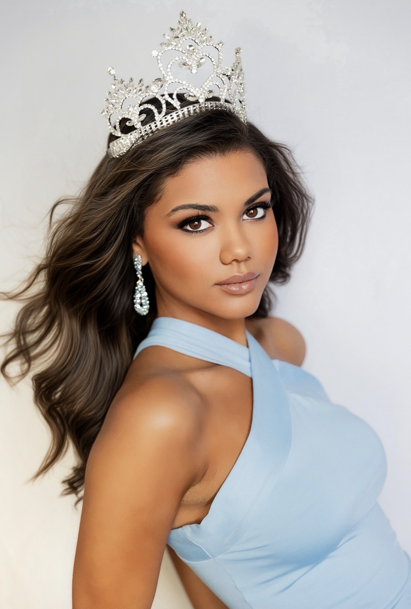 Woman wearing a crown and earrings, dressed in a light blue gown, with long wavy hair, looking at the camera.
