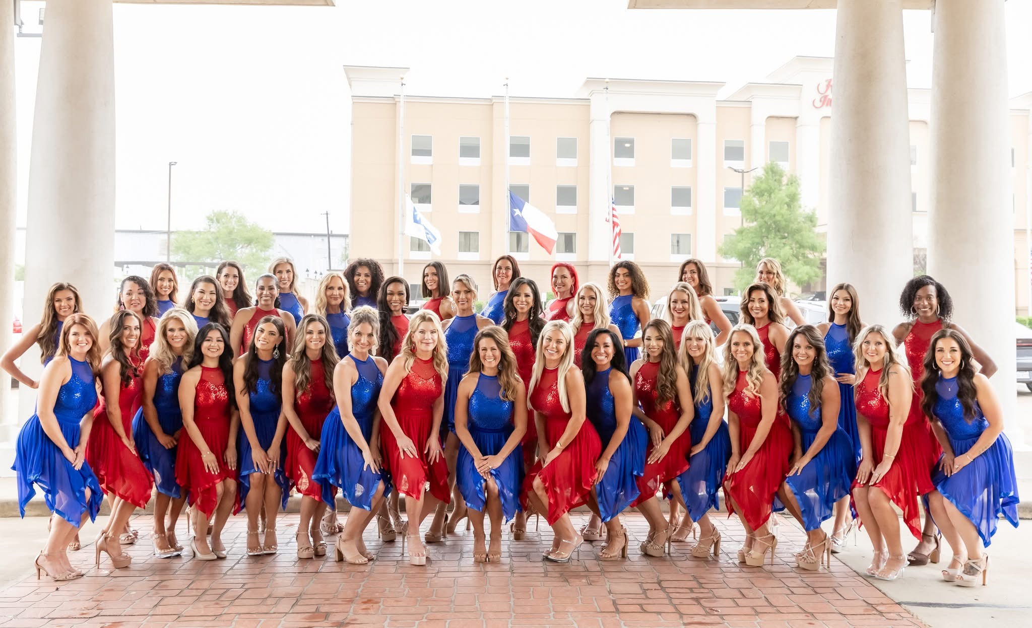 Group of women in red and blue dresses posing outdoors on a covered terrace, with buildings and trees in background.