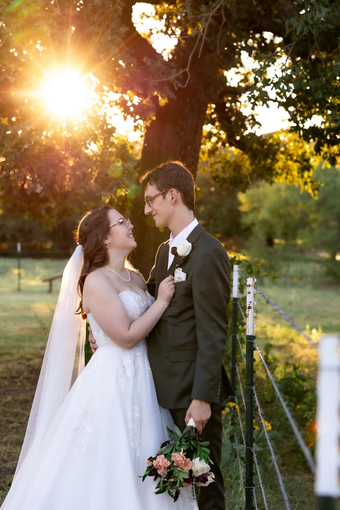 Bride and groom stand close together outdoors, sunlight shining through trees, holding flowers, gazing at each other.