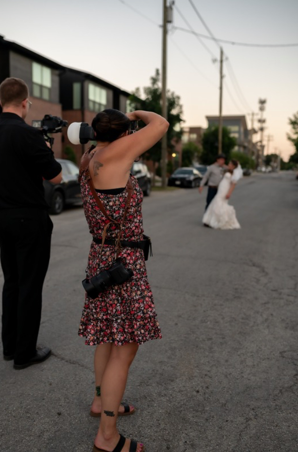 Photographer with camera on shoulder capturing a bride in a wedding dress walking away, on a street.